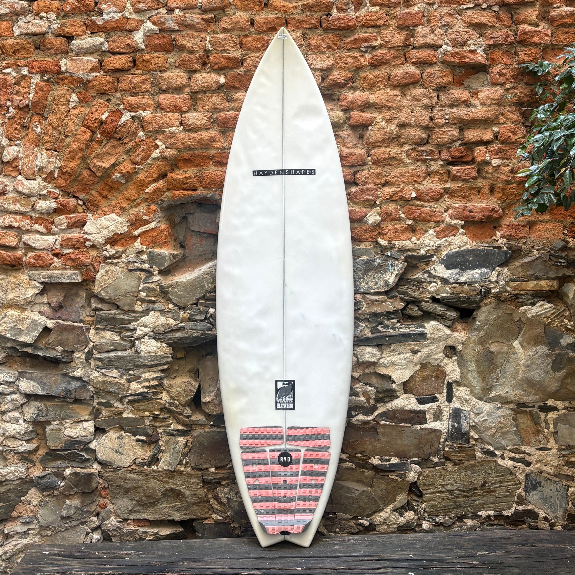 A white surfboard with a red and black striped tail patch, leaning against a rustic brick and stone wall.