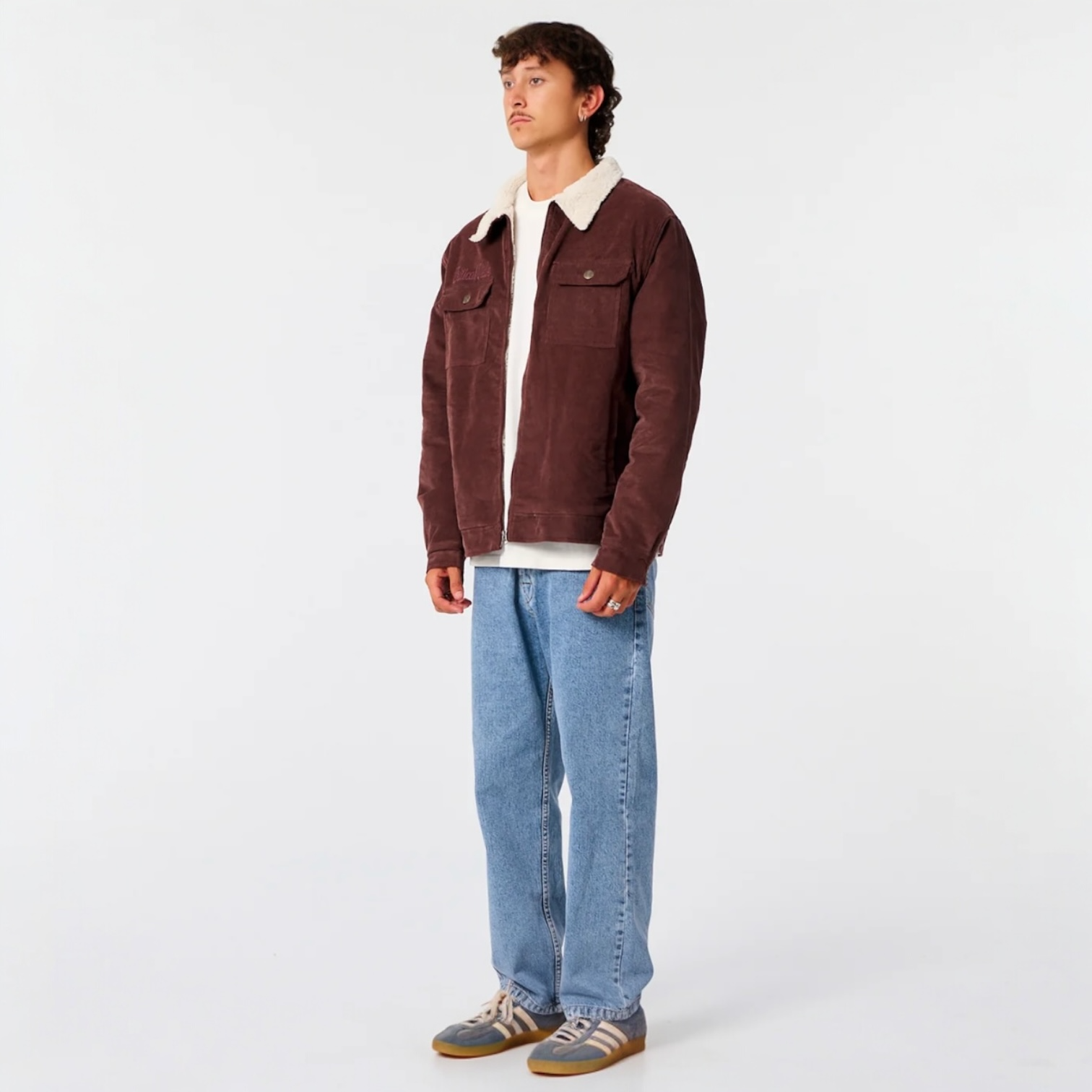 Man wearing a brown jacket with a white collar, white shirt, blue jeans, and brown shoes on a white background