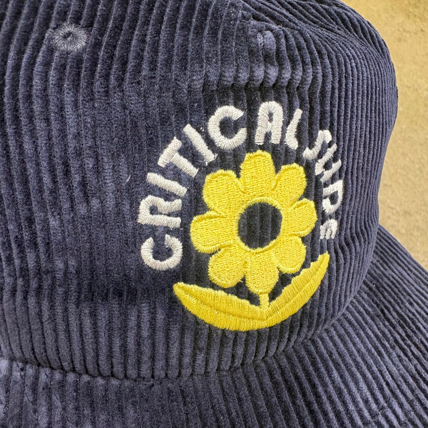 Gray corduroy jacket with a yellow flower and 'Critical Huds' logo on a beige background