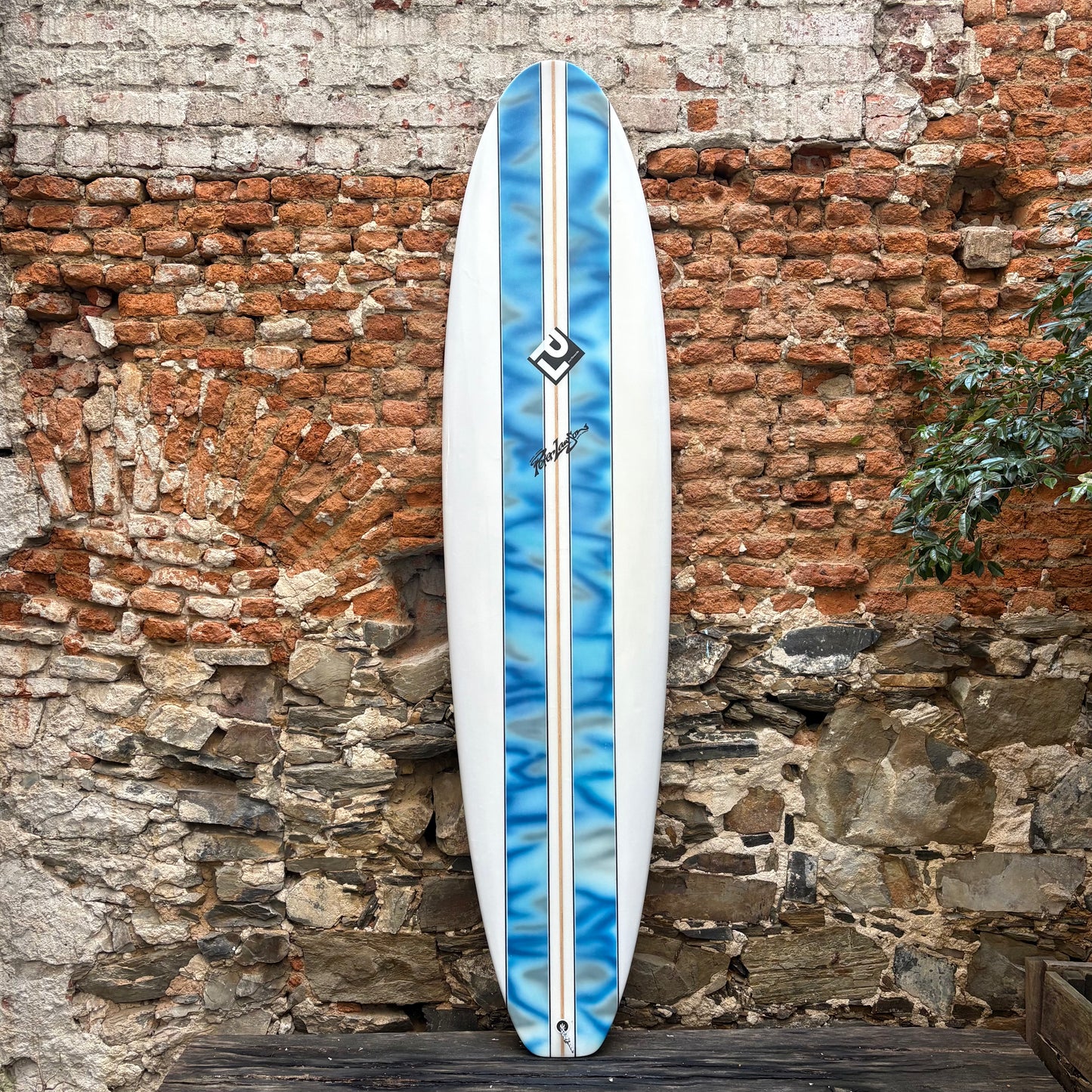 Blue and white surfboard with a logo against a rustic stone wall.