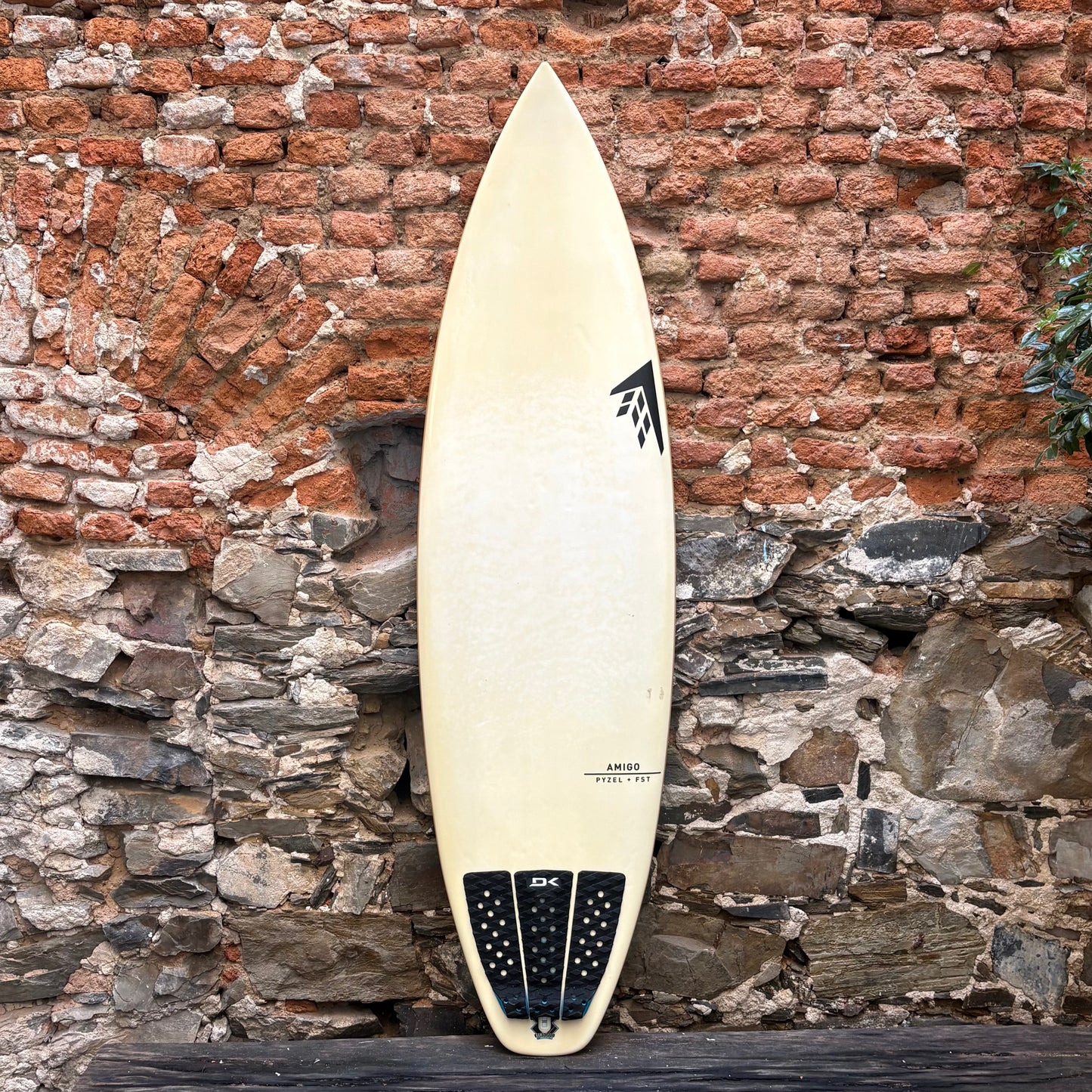 White surfboard with black logo against a rustic stone and brick wall.