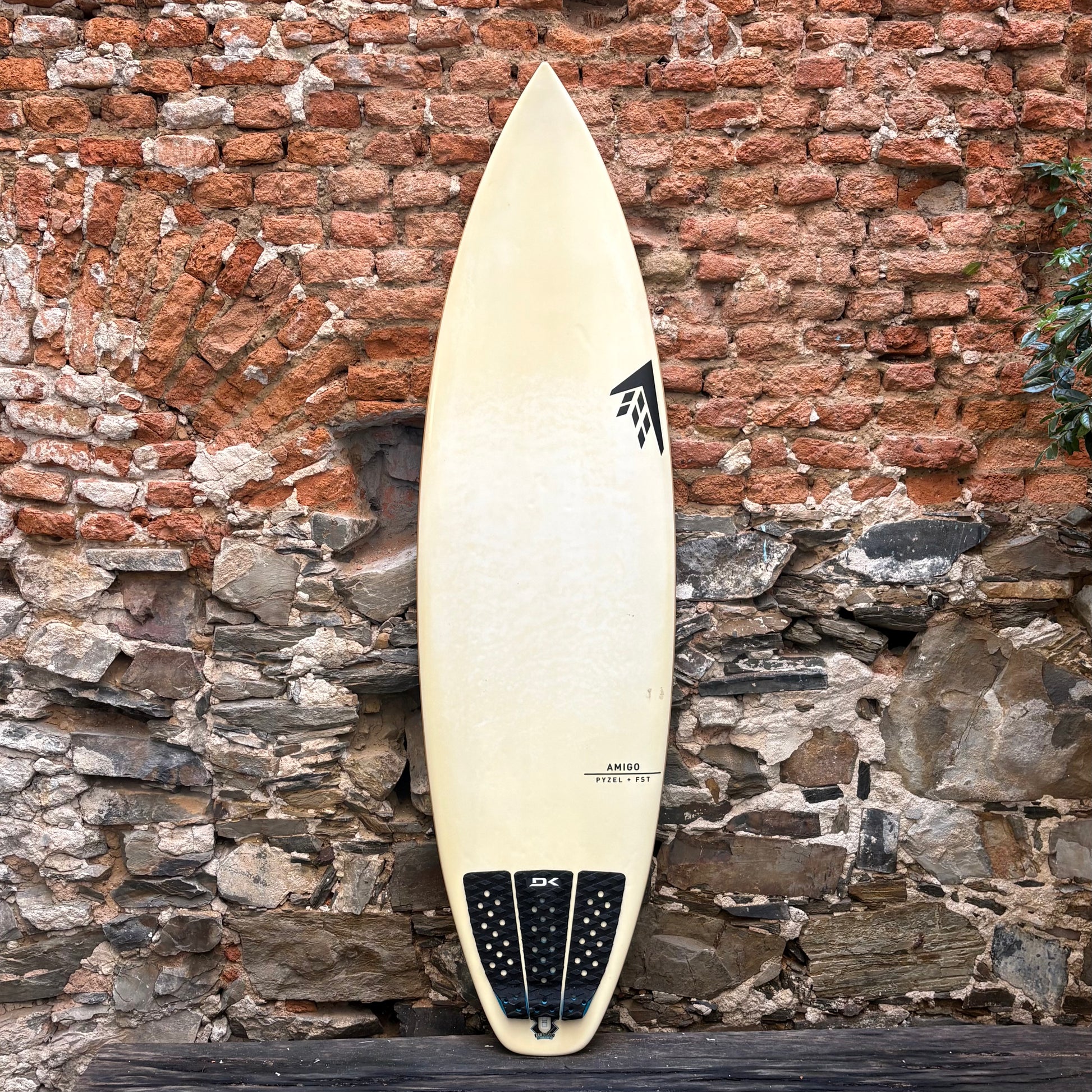 White surfboard with black logo against a rustic stone and brick wall.