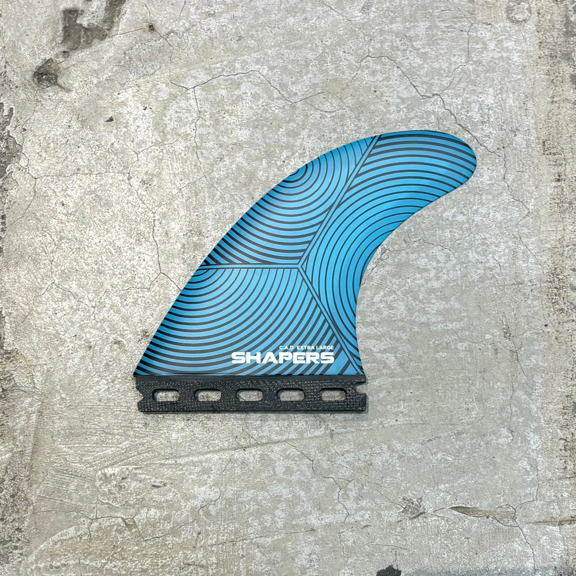 A blue and black surfboard fin with a geometric wave pattern and the brand name 'SHAPERS' printed on it, placed on a textured concrete surface.