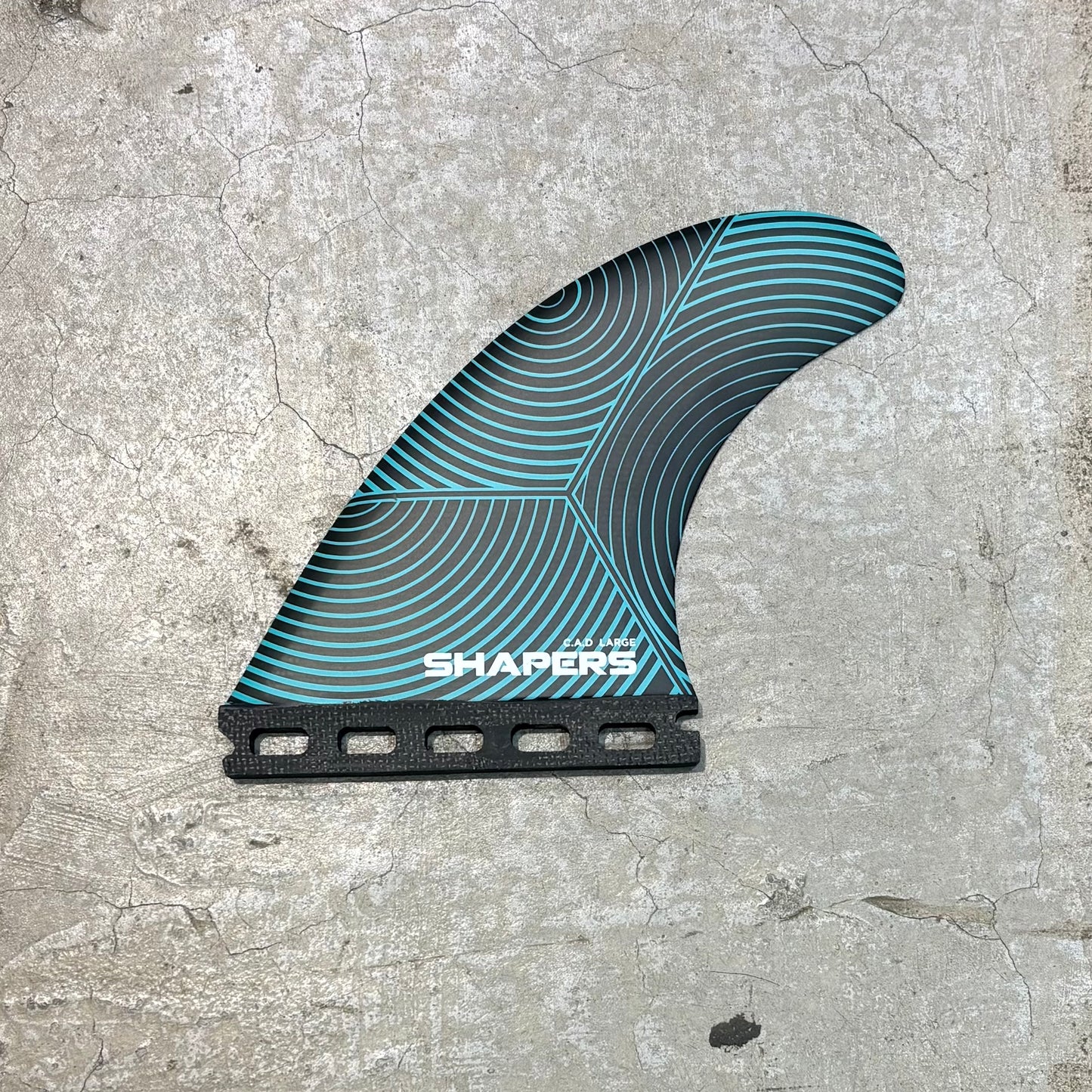 A surfboard fin blade with a black and teal wave pattern, featuring the brand name 'SHAPERS' in white on the base, set against a textured concrete background.