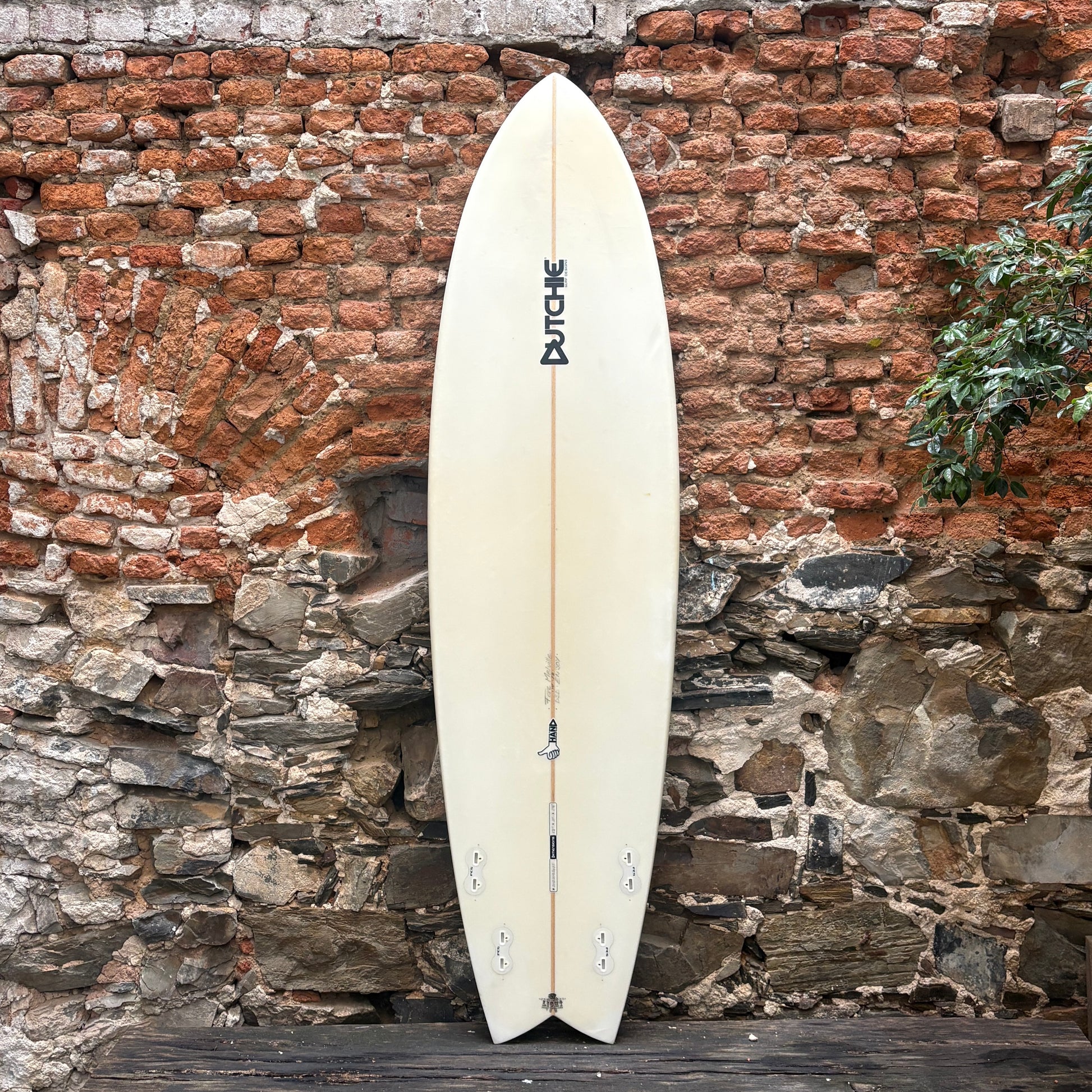 White surfboard with a brand logo against a rustic stone wall.