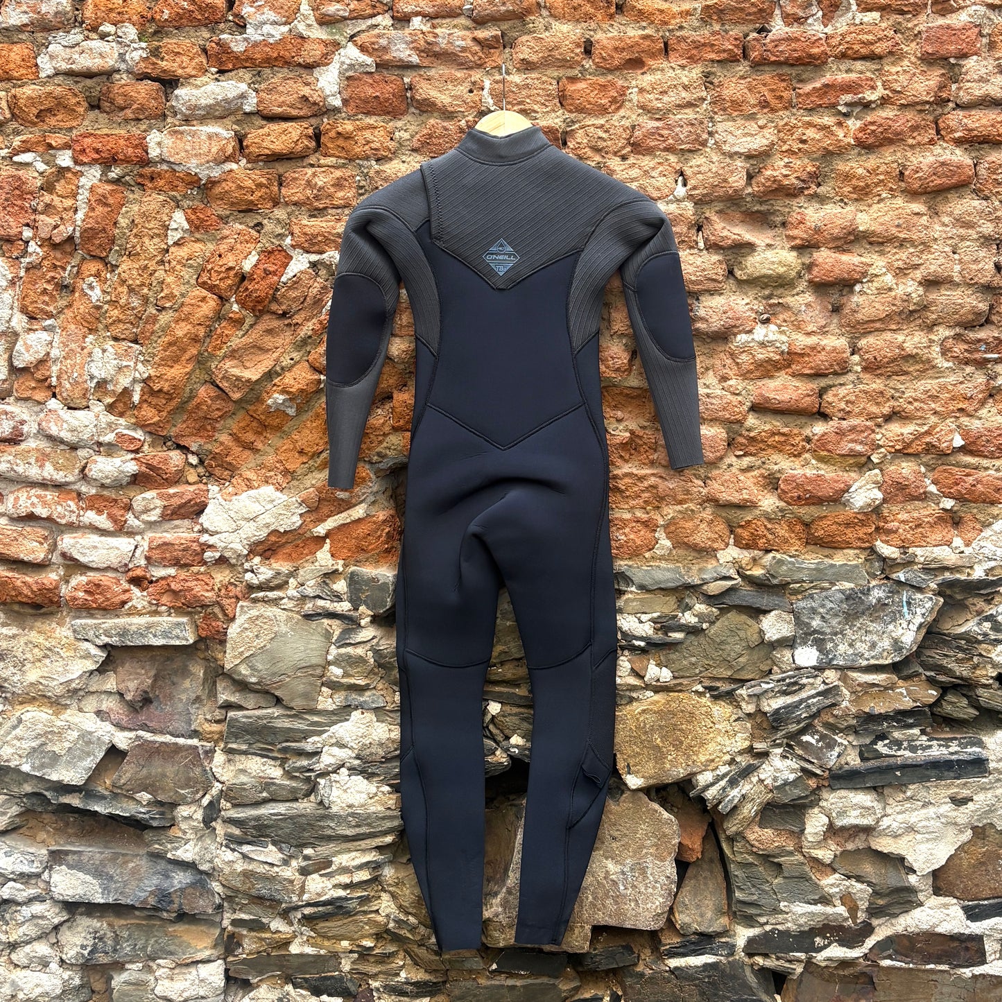 Back view of O'Neill Youth Hyperfreak 4/3mm+ Chest Zip Wetsuit - Black/Tempered Steel in front of rustic stone wall