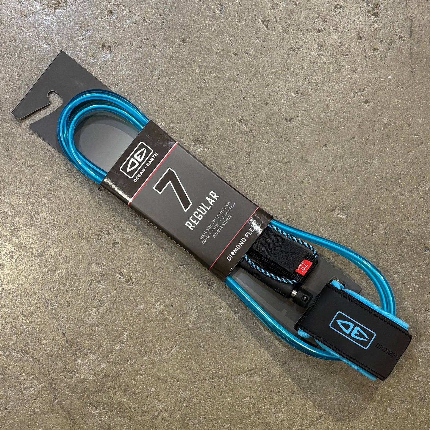 A blue 7 Regular surf leash with its retail packaging, placed diagonally on a textured gray concrete floor.