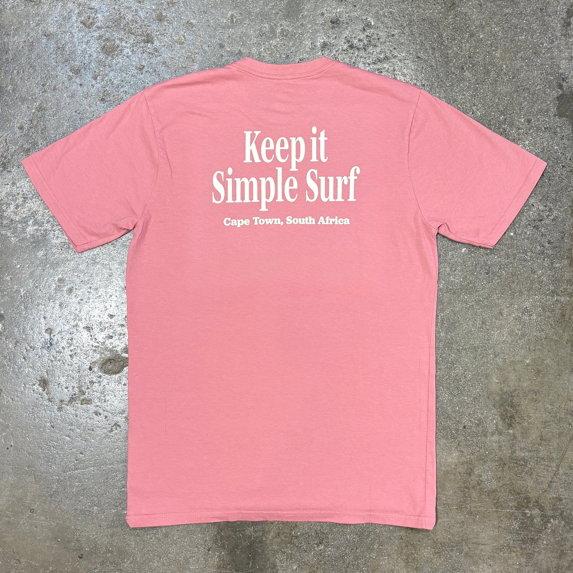 A pink short-sleeve t-shirt laid flat on a textured gray concrete surface, featuring the white printed text 'Keep it Simple Surf' and 'Cape Town, South Africa' on the back.