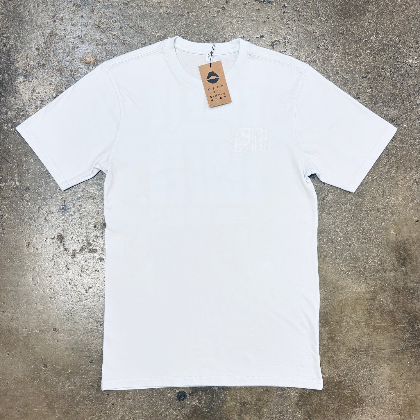 A sky blue short-sleeve crewneck t-shirt with a small tonal chest logo, laid flat on a textured concrete floor. A brown paper hangtag is attached to the collar.
