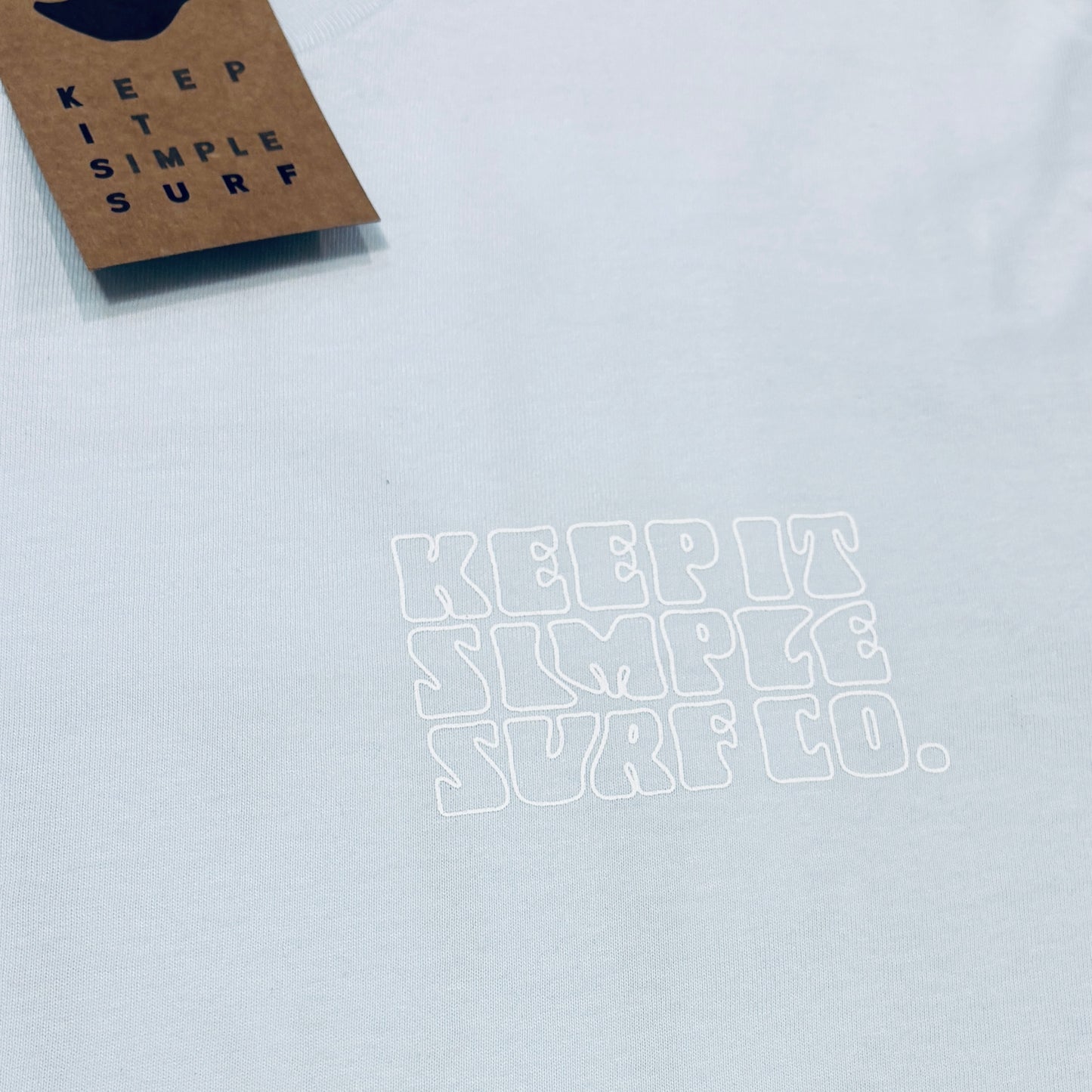 A close-up of a sky blue T-shirt or sweatshirt fabric with a large outlined 'KEEP IT SIMPLE SURF CO.' logo printed in white on the front.