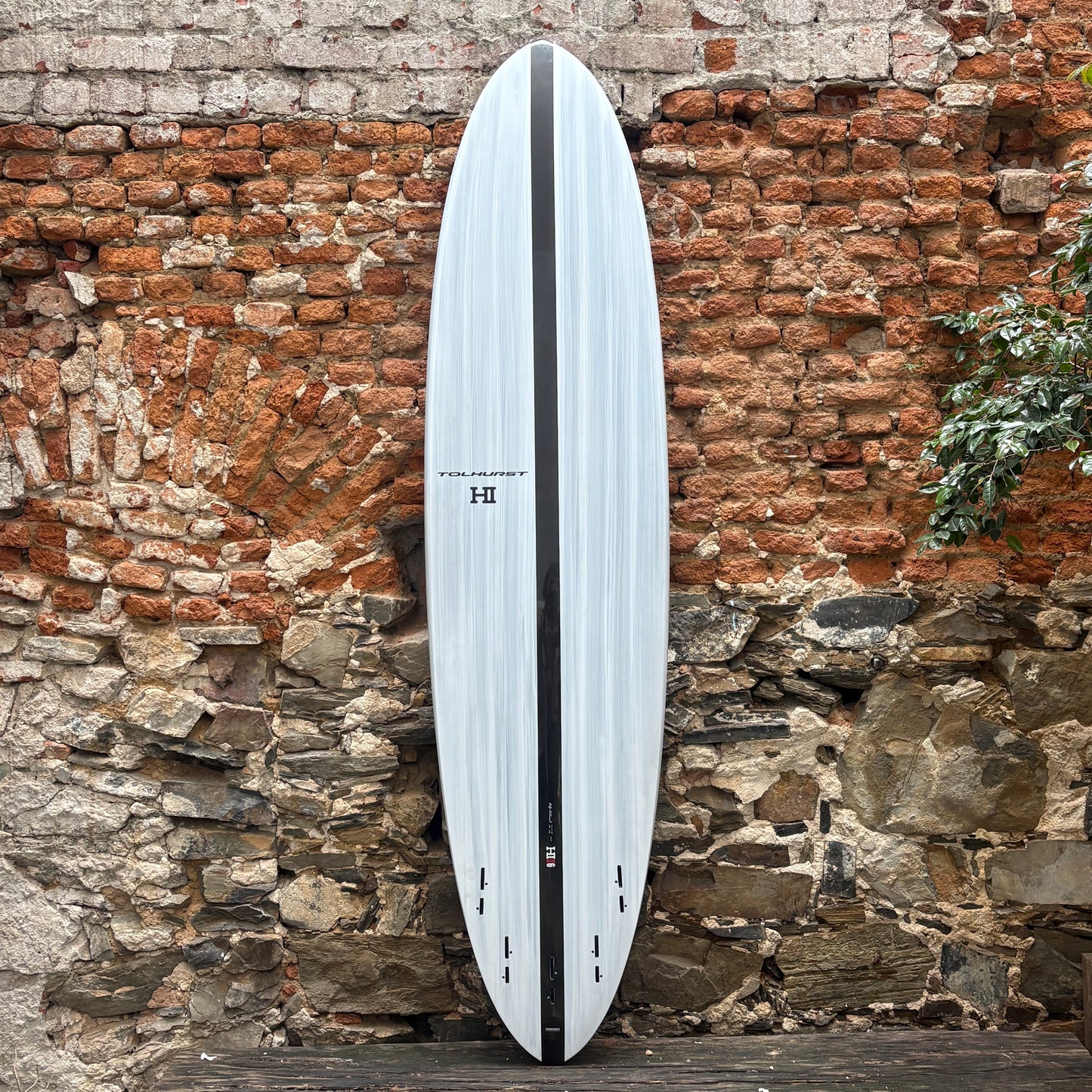 A long surfboard with a white and light gray woodgrain pattern and a black center stripe, leaning against a rustic brick and stone wall.