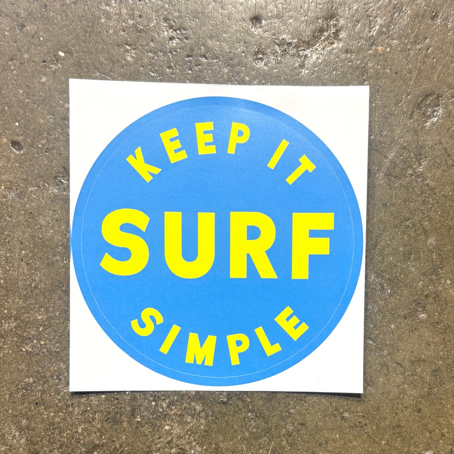 A square white sticker with a blue circle in the center featuring the yellow text 'KEEP IT SIMPLE SURF', placed on a textured gray concrete surface.