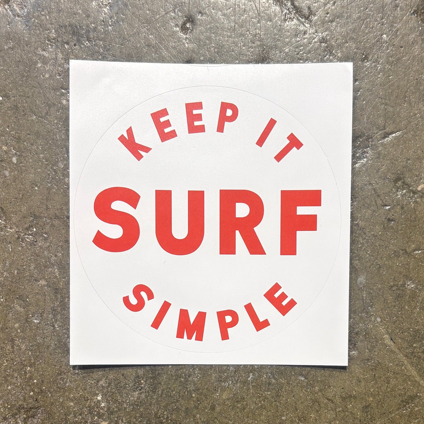 A square white sticker with a circular design featuring the words 'KEEP IT SIMPLE SURF' in red lettering, placed on a textured gray concrete surface.