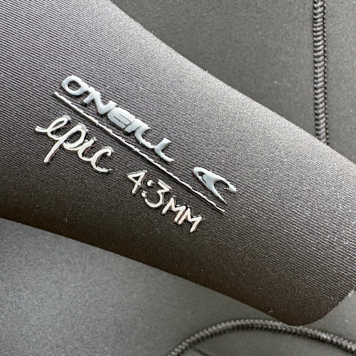 Close-up of a dark gray nwetsuit sleeve with silver foil branding that reads 'ONEILL epic 4:3MM' on textured neoprene fabric.