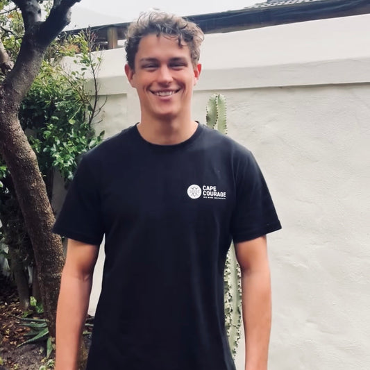 A smiling young man wearing a black short-sleeve t-shirt with a white 'Cape Town University' logo on the left chest, standing outdoors in front of a white wall and greenery.