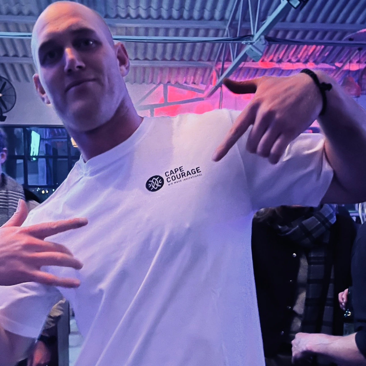 A man wearing a white t-shirt with a black 'CAPE COURAGE' logo on the left chest, posing in a dimly lit indoor venue with colored ambient lighting.