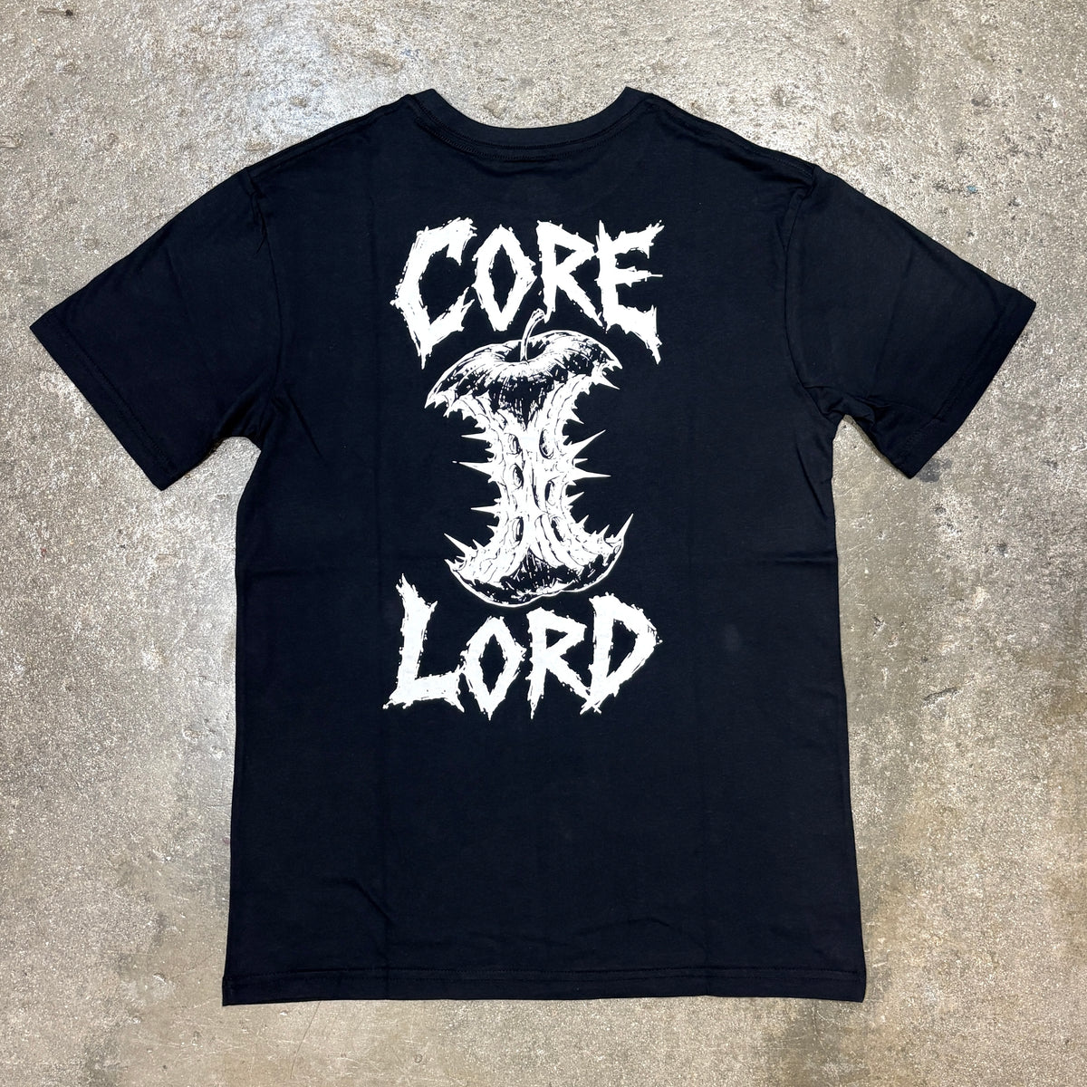A black short-sleeve t-shirt laid flat on a textured concrete floor, featuring a large white graphic print on the back with the words 'CORE LORD' and a stylized thorn-like symbol.