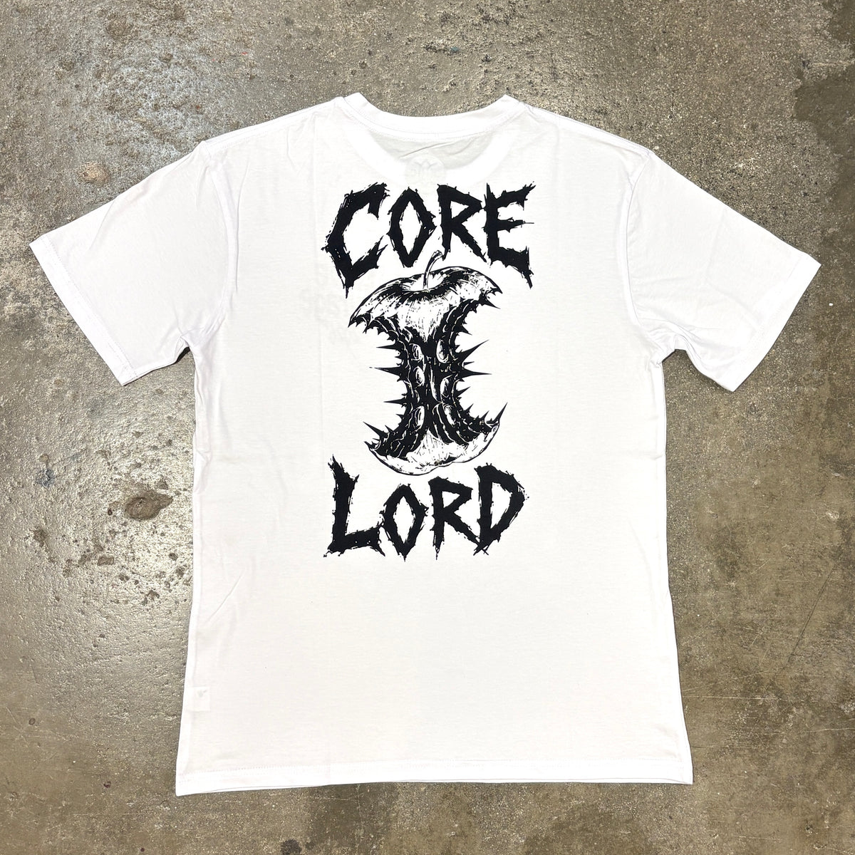 A white short-sleeve t-shirt laid flat on a textured concrete floor, featuring a large black graphic print on the front with the words 'CORE' and 'LORD' framing a stylized thorn-like emblem.
