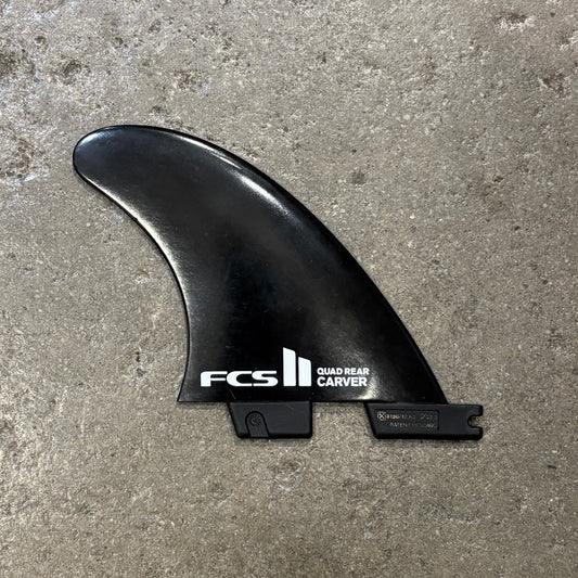A black FCS Quad Rear Carver surfboard fin resting on a textured concrete floor. The fin features white printed branding and specifications.