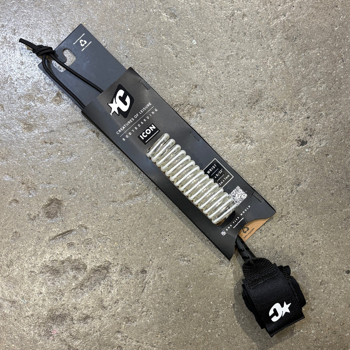 A white bodyboard leash with a white coiled leash line and a black wrist strap, shown attached to its black retail packaging card on a gray concrete floor. The packaging features the XC Surf brand logo and the model name ICON.