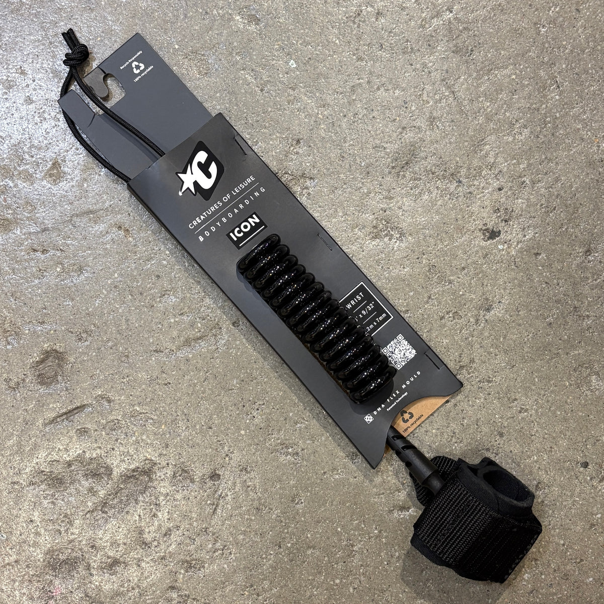 A black coiled leash with a black wrist strap attached, mounted on a branded black retail card and photographed on a concrete floor. The card shows the XC brand and the model name ICON.