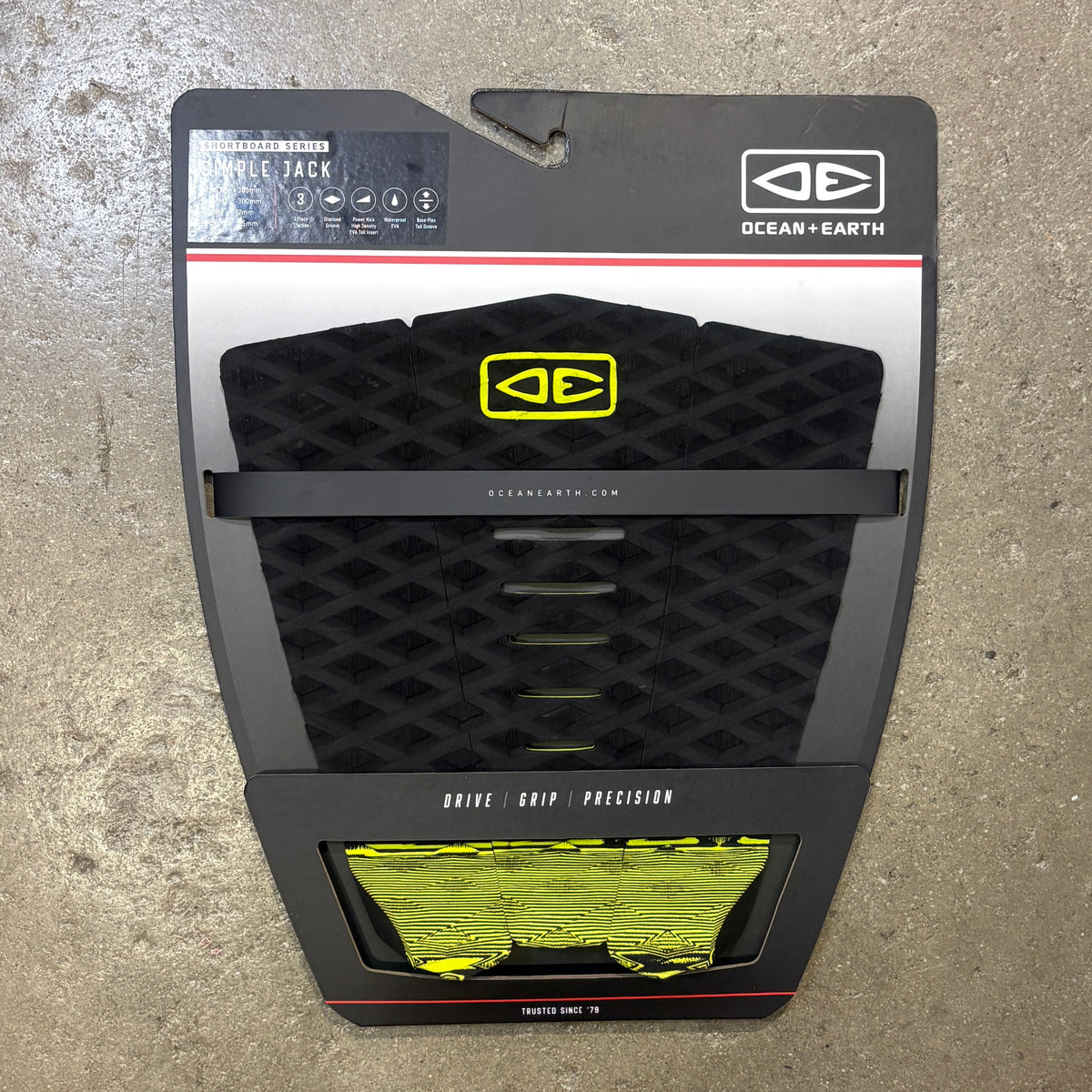 Ocean Earth Simple Jack Traction Deck in its original retail packaging, featuring a black textured grip pad with a yellow logo and a clear window showing the yellow and black grip tape underneath, set against a concrete floor background.