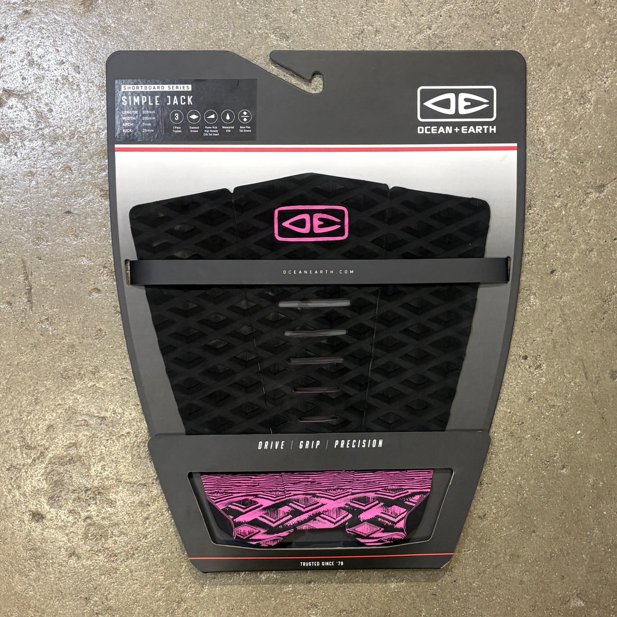 Ocean Earth Simple Jack traction pad in black with bright pink graphics, shown in its retail packaging against a concrete floor background.