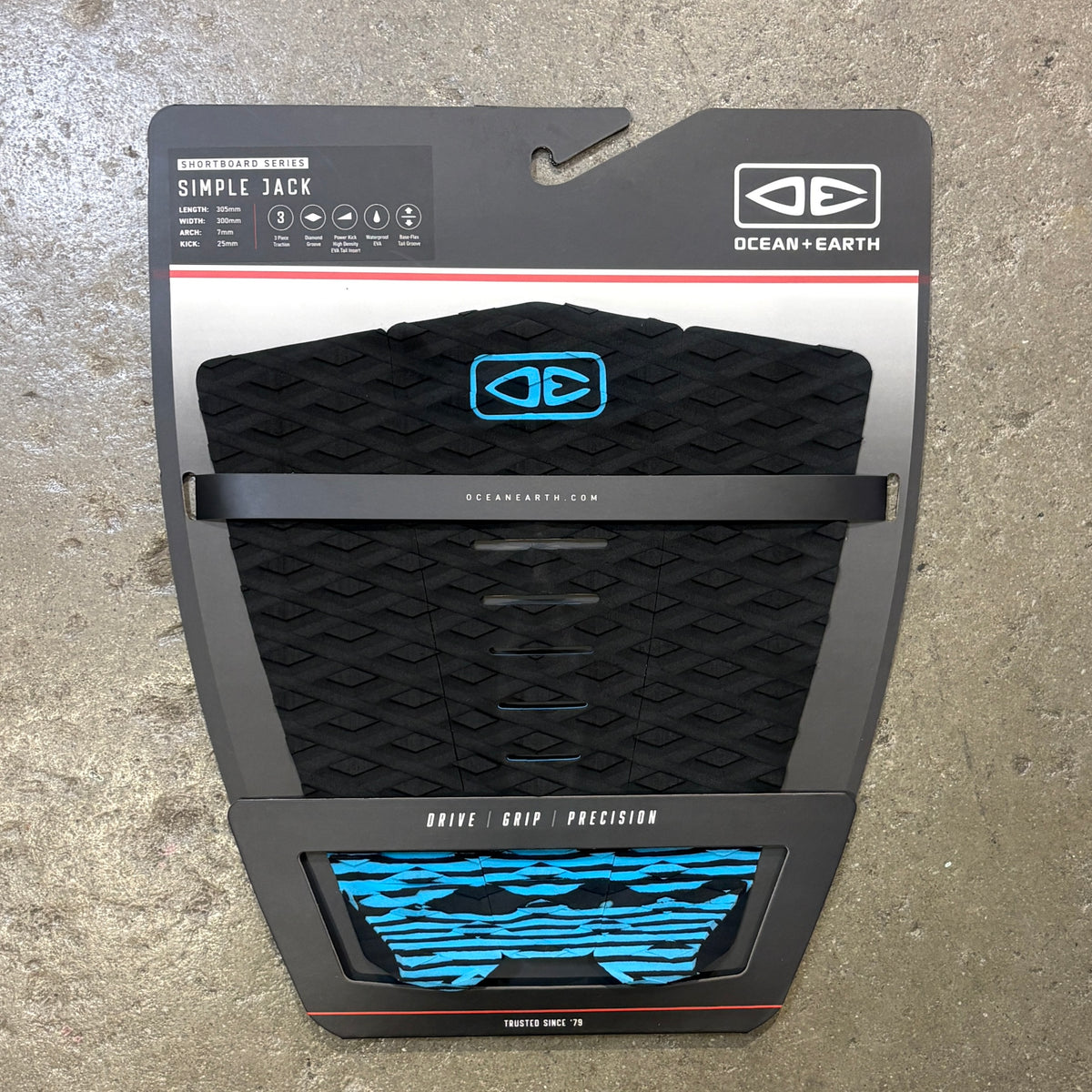 Ocean Earth Simple Jack traction pad in black with bright blue accents, shown in its original retail packaging against a concrete floor background.