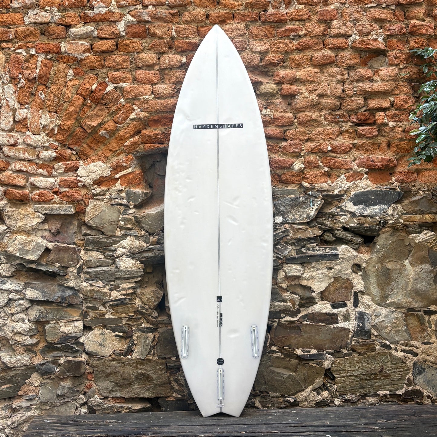 A white surfboard standing upright against a rustic brick and stone wall.