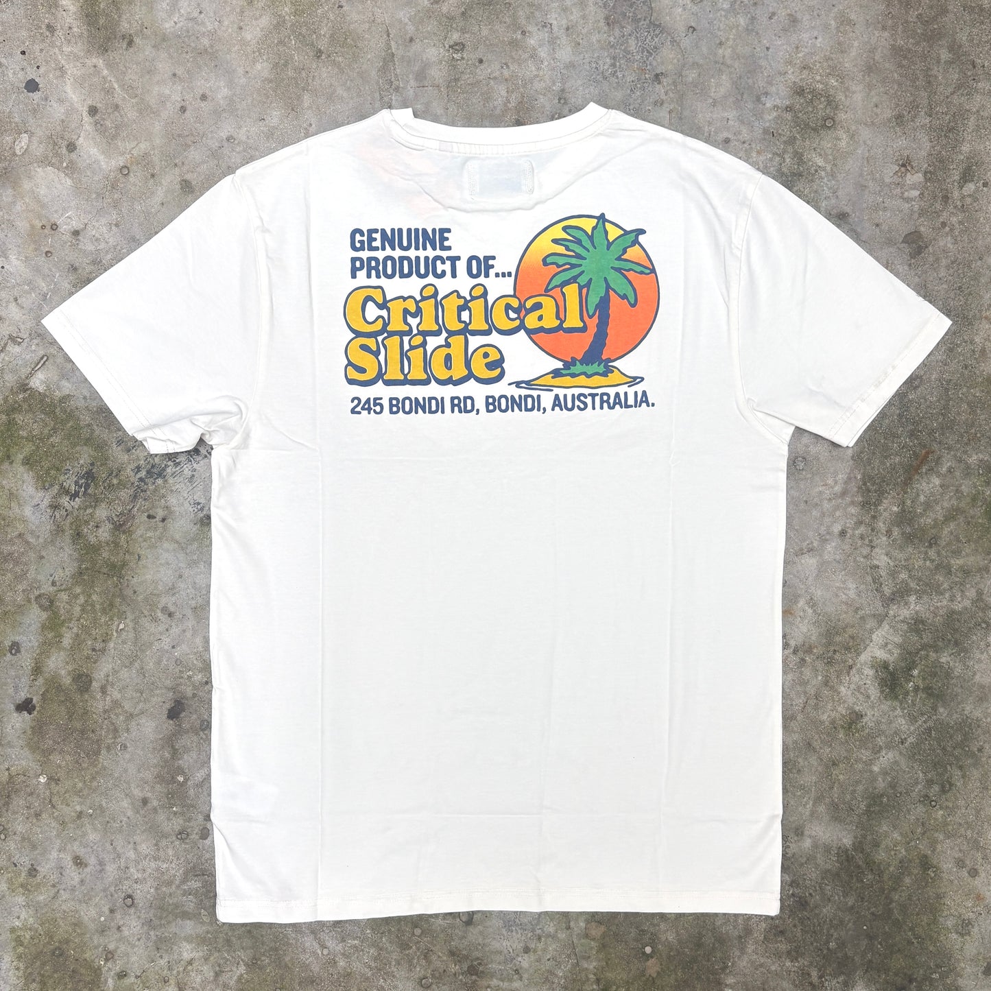 A white short-sleeve t-shirt laid flat on a textured gray concrete surface, featuring a colorful graphic print on the chest with a palm tree, sunset, and the text 'Genuine Product of Critical Slide'.