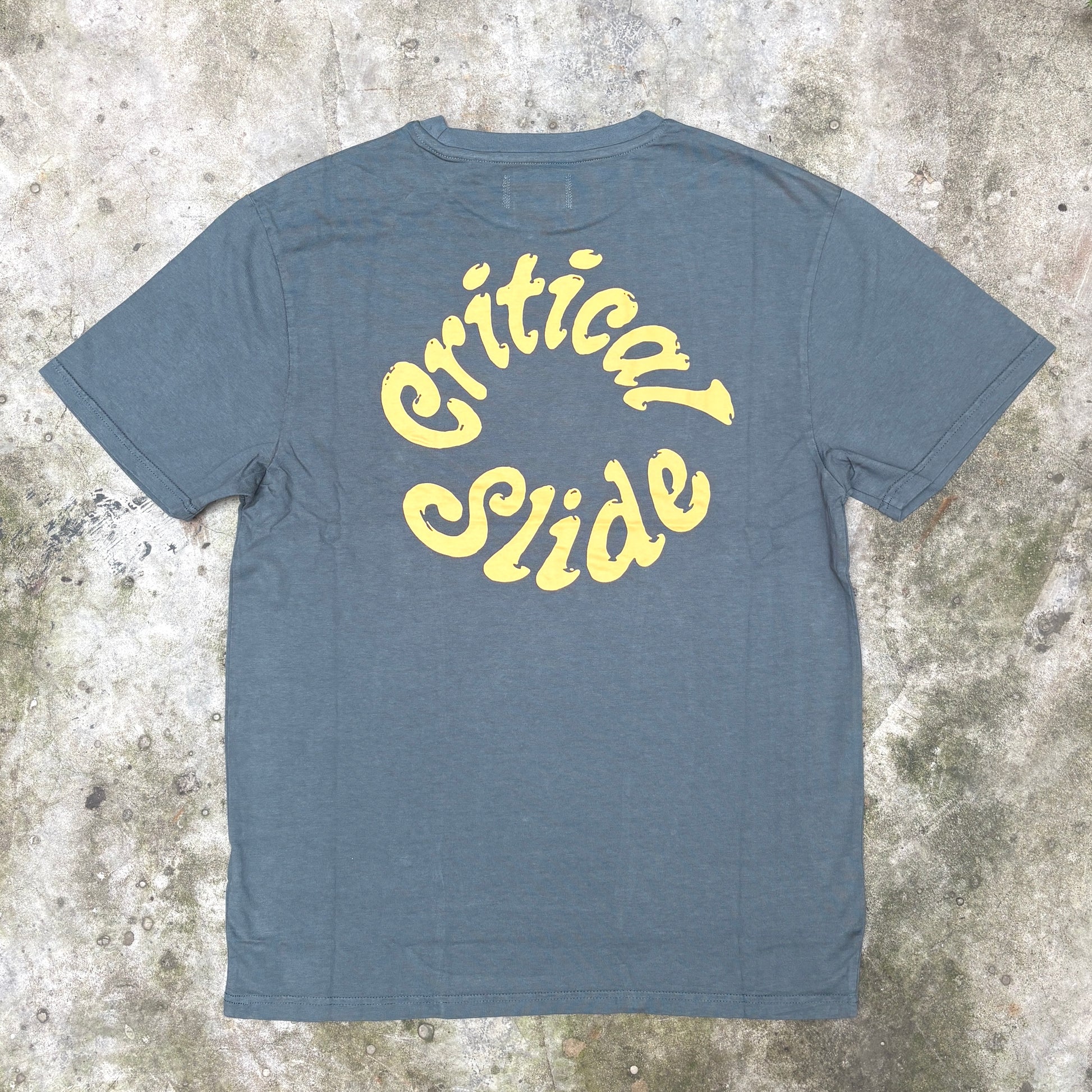 A slate blue heather t-shirt with 'Critical Slide' printed in yellow across the chest, laid flat on a textured concrete surface.
