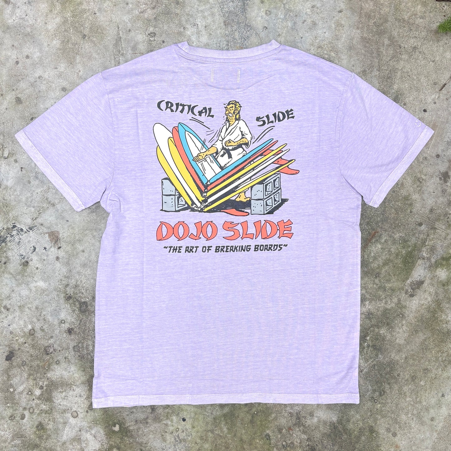 A light lavender heather t-shirt featuring a retro graphic of a surfer riding a slide with the text 'CRITICAL SLIDE' and 'DOJO SLIDE' on a concrete background.