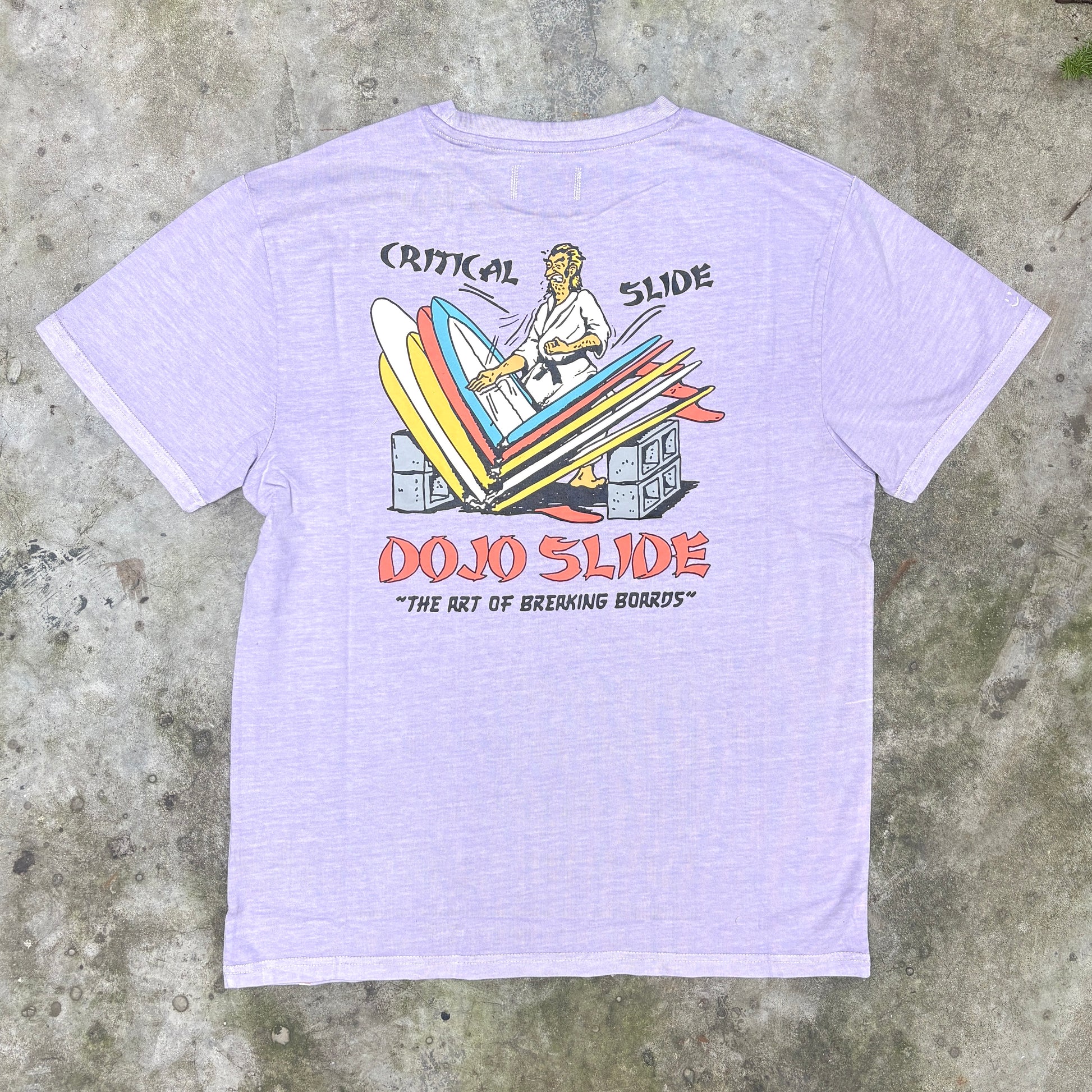 A light lavender heather t-shirt featuring a retro graphic of a surfer riding a slide with the text 'CRITICAL SLIDE' and 'DOJO SLIDE' on a concrete background.