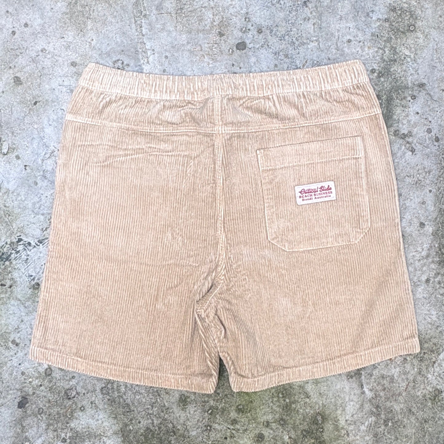 A pair of light tan corduroy shorts shown from the back, featuring an elastic waistband, a back pocket with a red and white 'Vintage Rock' label, and a ribbed hem, laid flat on a gray concrete surface.