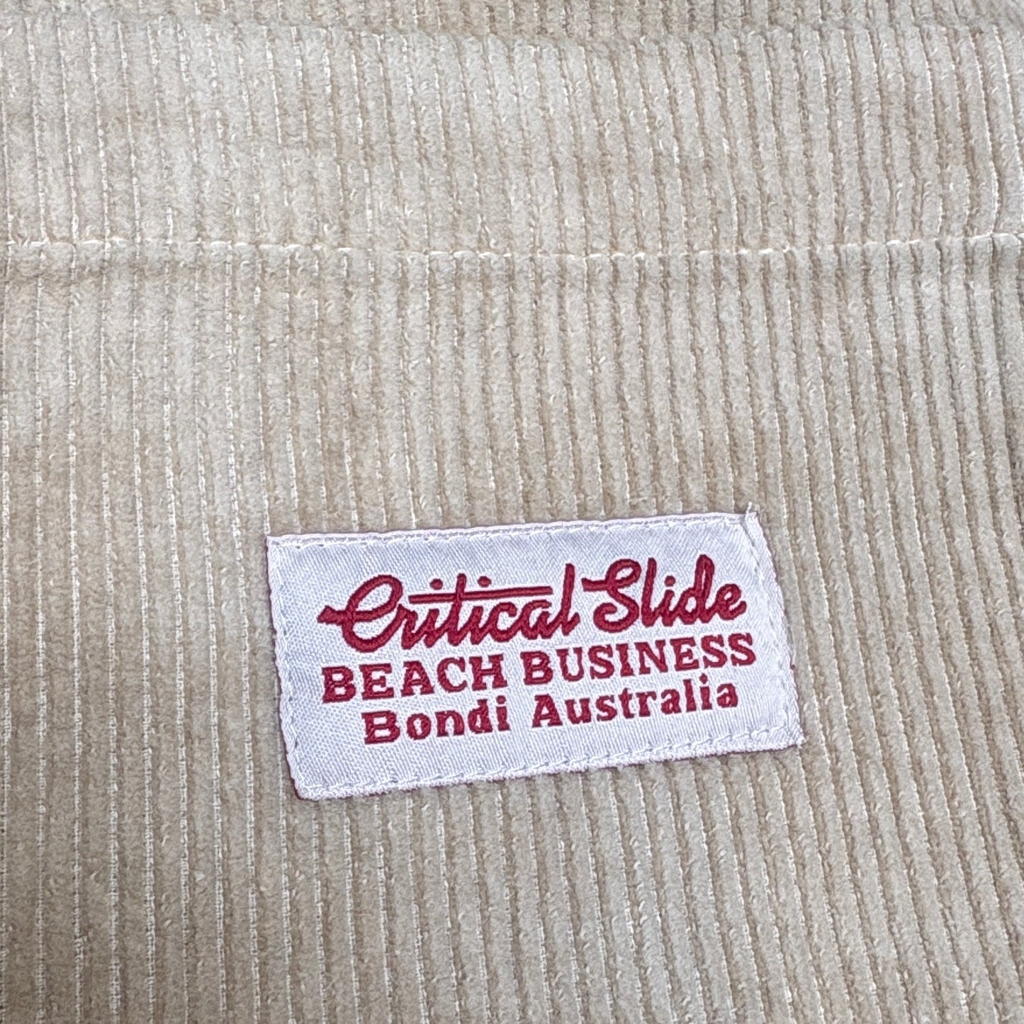 Close-up of a beige ribbed fabric label reading 'Critical Slide Beach Business Bondi Australia' in red text on a white patch.