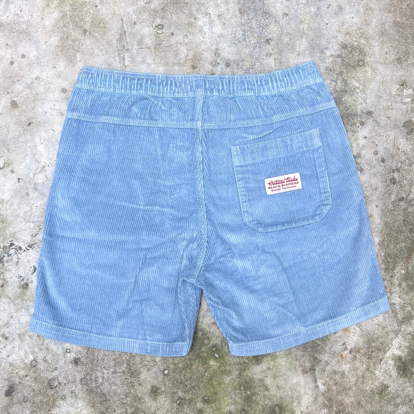 Light blue corduroy shorts with an elastic waistband and a back pocket featuring a red and white 'Hastings Brand' label, laid flat on a textured concrete surface.