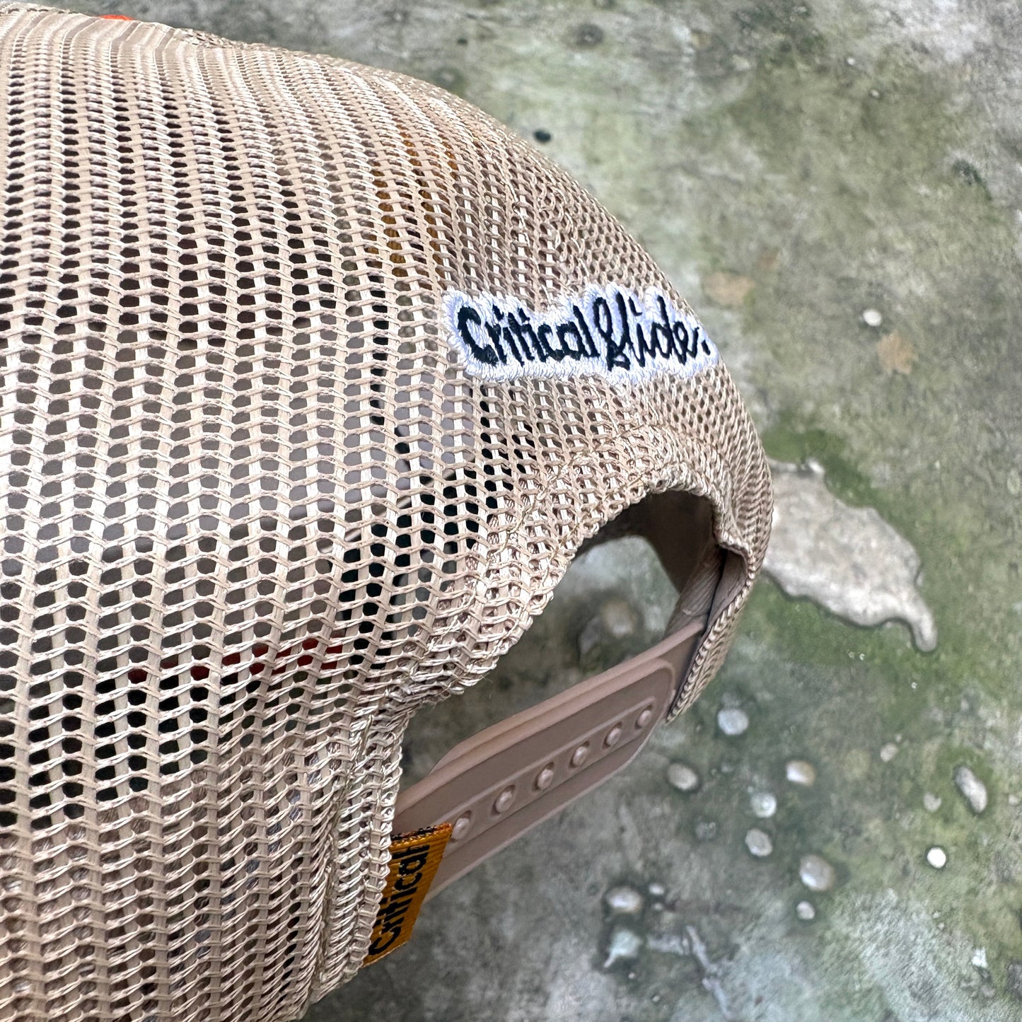 A close-up of a beige mesh trucker hat with a white 'Critical Kidz' sticker on the side, resting on a wet concrete surface.