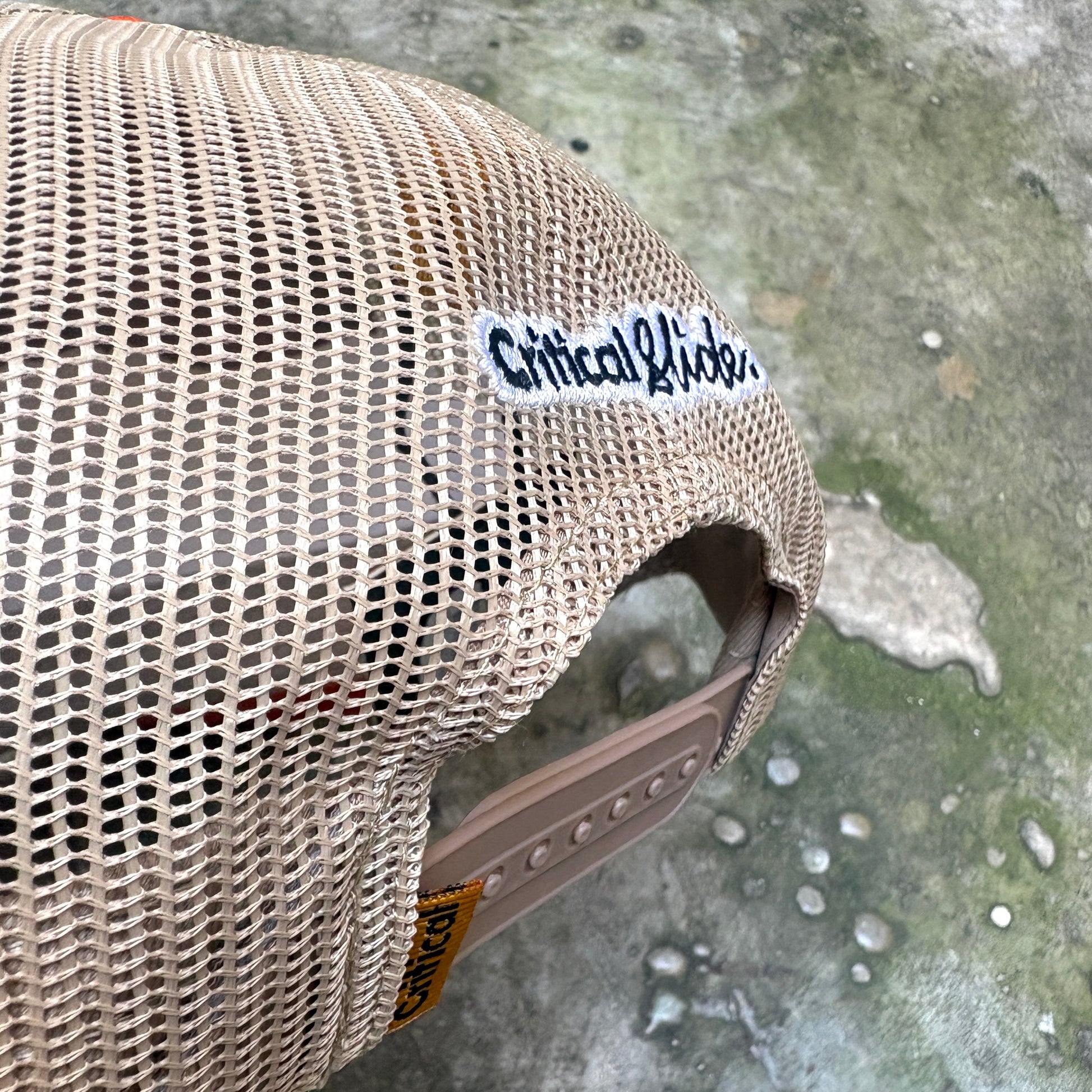 A close-up of a beige mesh trucker hat with a white 'Critical Kidz' sticker on the side, resting on a wet concrete surface.