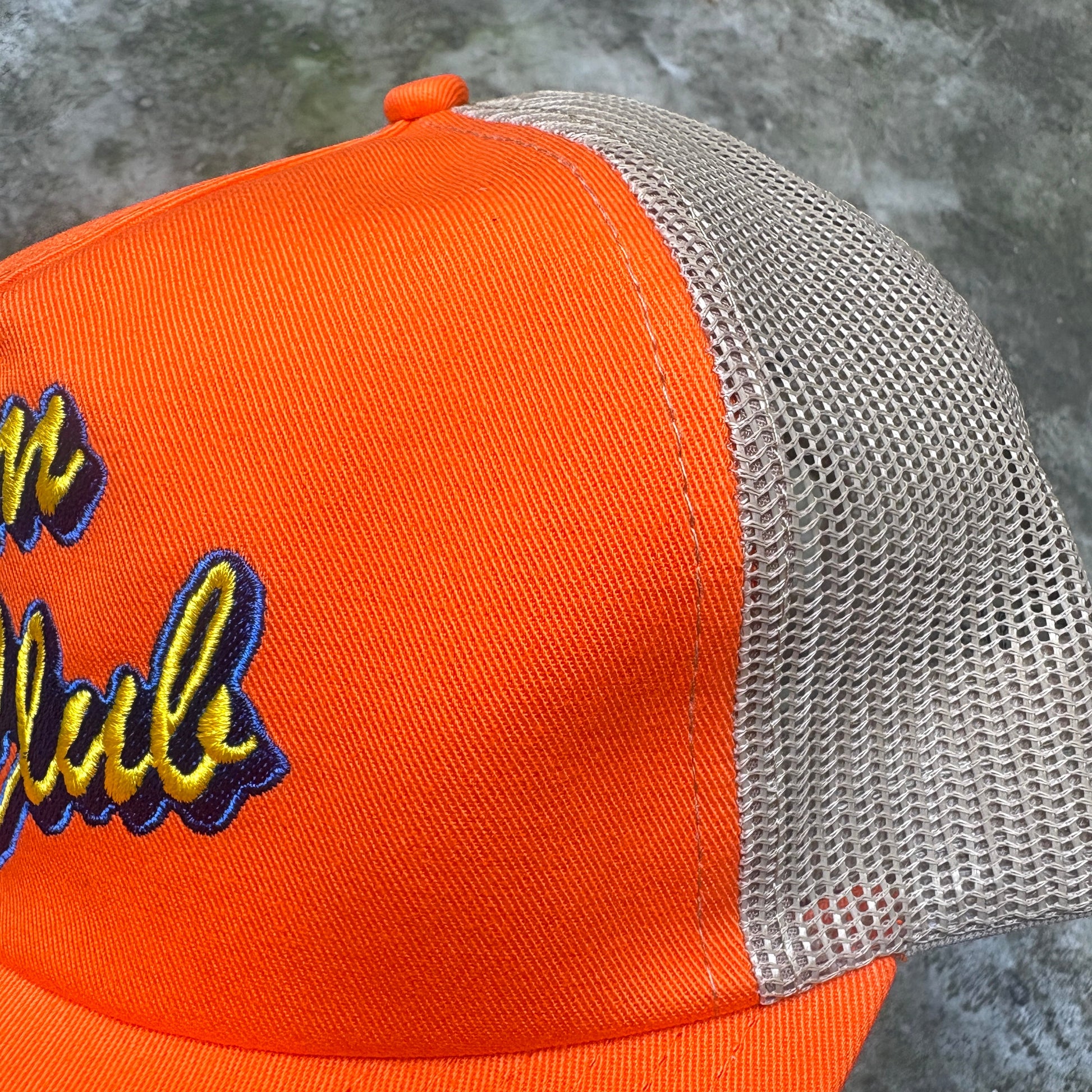 Close-up of an orange trucker cap with a beige mesh back and embroidered yellow and blue lettering on the front panel, photographed against a gray concrete surface.