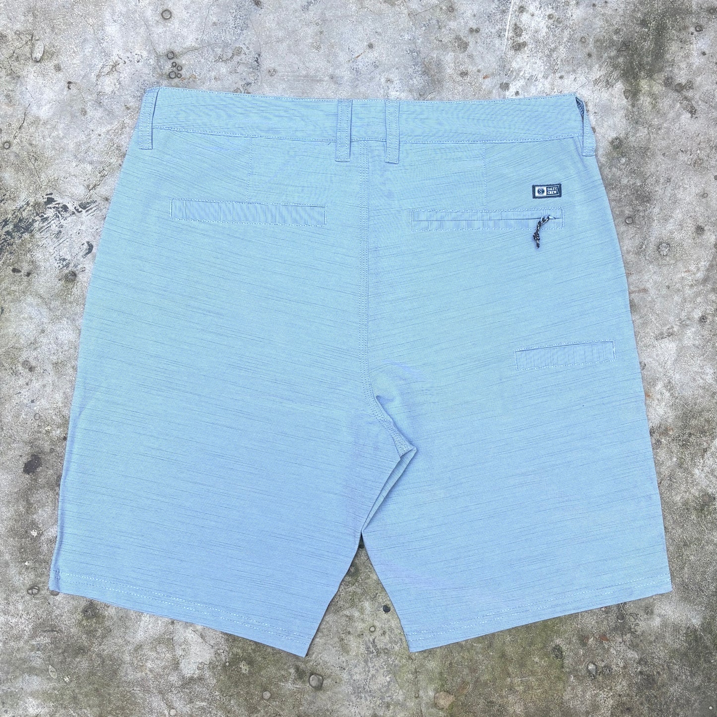 Light blue textured shorts shown from the back, laid flat on a concrete surface. The shorts feature belt loops, a button closure, two back welt pockets, and a small black brand patch on the upper right side.