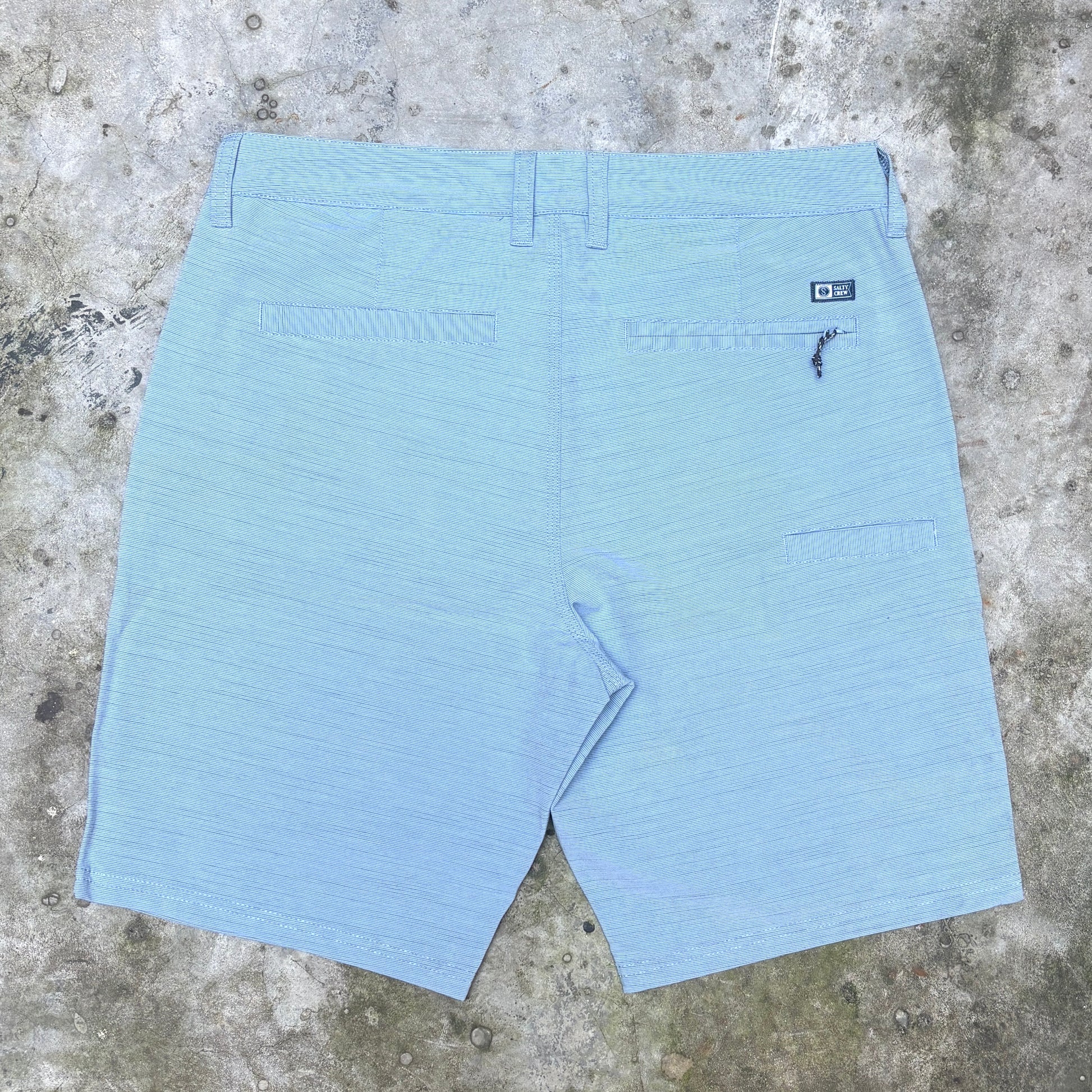 Light blue textured shorts shown from the back, laid flat on a concrete surface. The shorts feature belt loops, a button closure, two back welt pockets, and a small black brand patch on the upper right side.