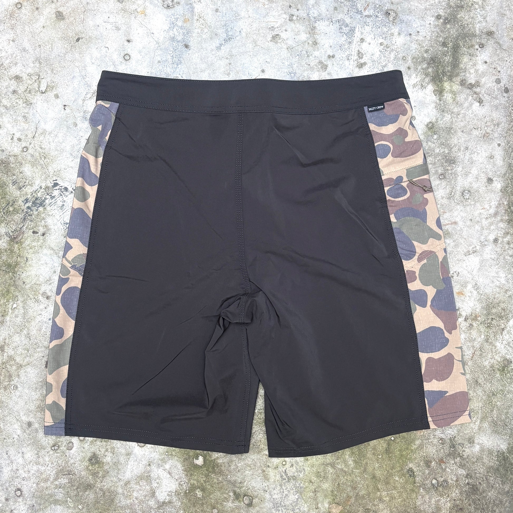 Black surf shorts with side panels in a camouflage pattern, laid flat on a textured concrete surface.