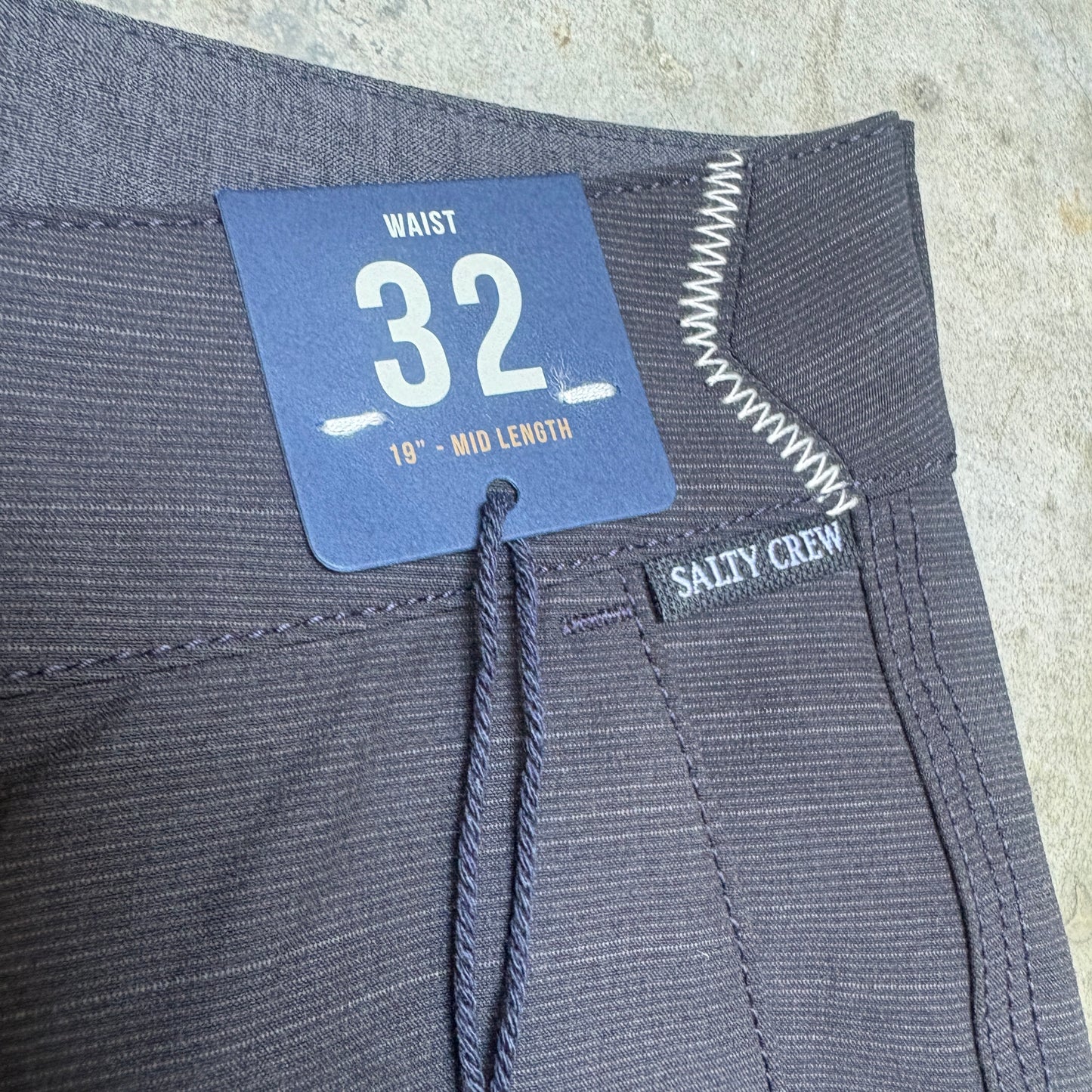 Close-up of dark gray striped pants showing the waistband, a blue size tag reading 32, and a small Salty Crew brand label.