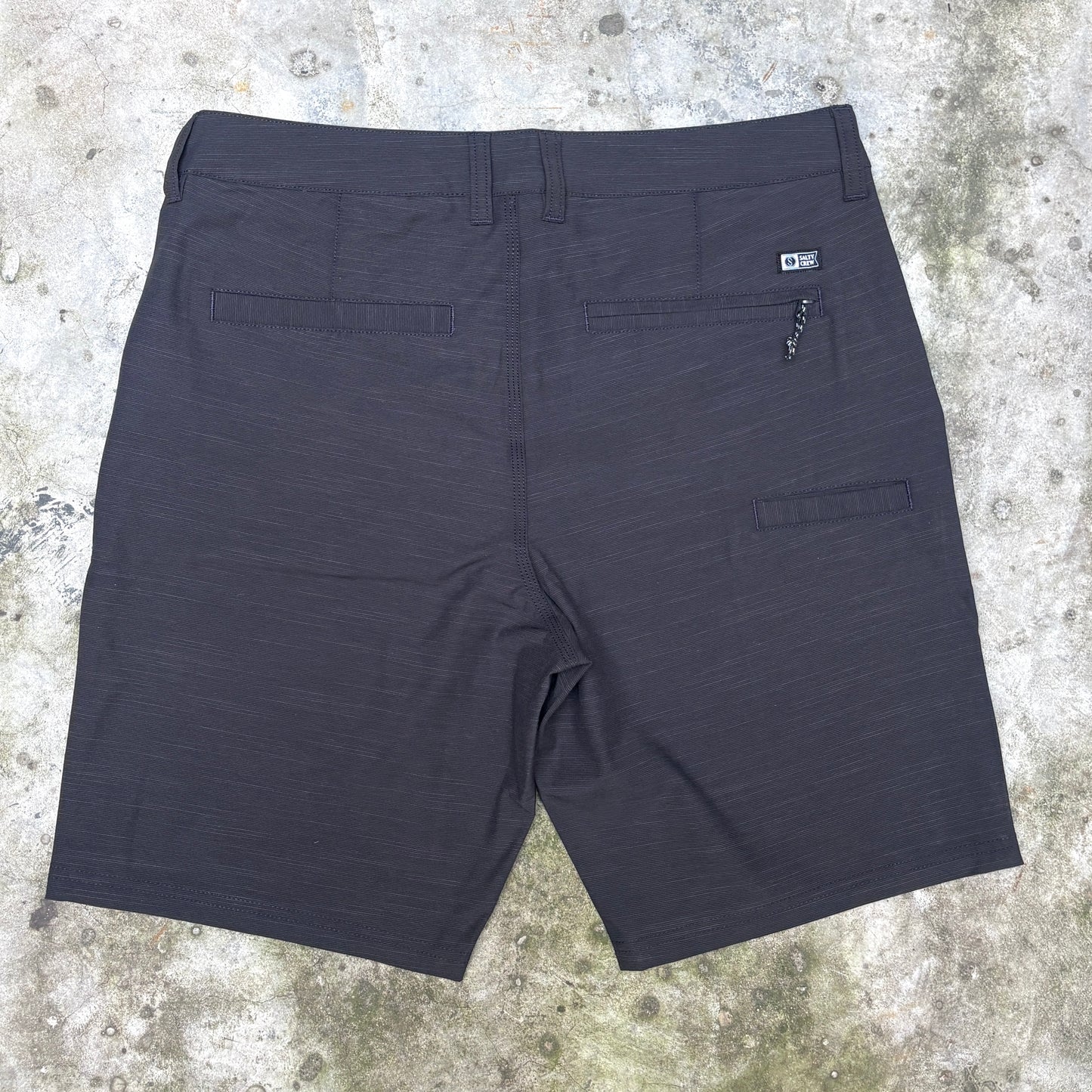 Dark charcoal gray textured athletic shorts shown from the back, laid flat on a concrete surface. Features include a waistband, two rear patch pockets, and a zippered pocket on the right side.