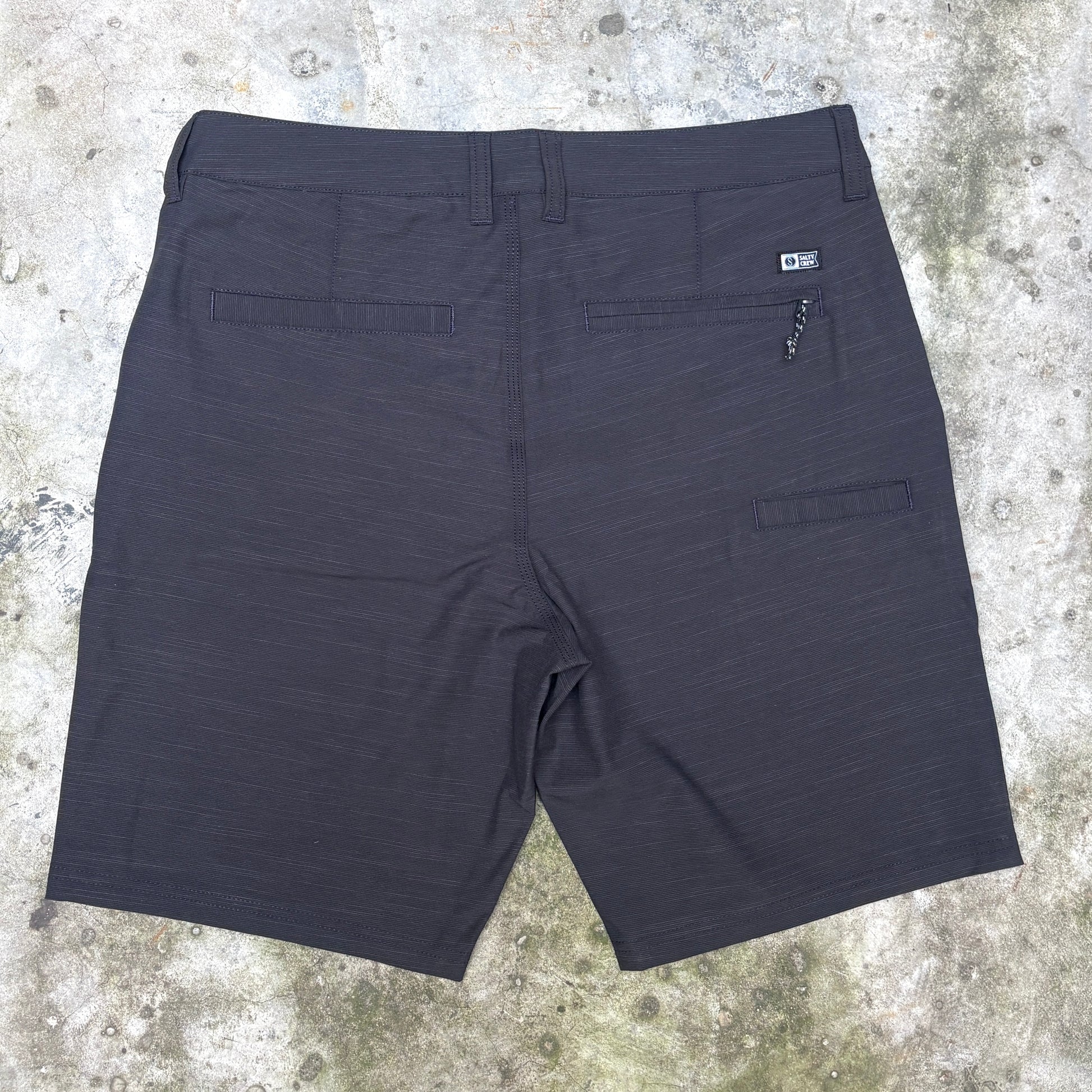 Dark charcoal gray textured athletic shorts shown from the back, laid flat on a concrete surface. Features include a waistband, two rear patch pockets, and a zippered pocket on the right side.