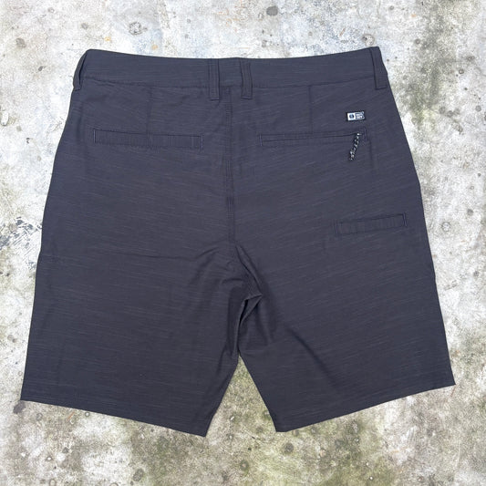 Dark charcoal gray textured athletic shorts shown from the back, laid flat on a concrete surface. Features include a waistband, two rear patch pockets, and a zippered pocket on the right side.