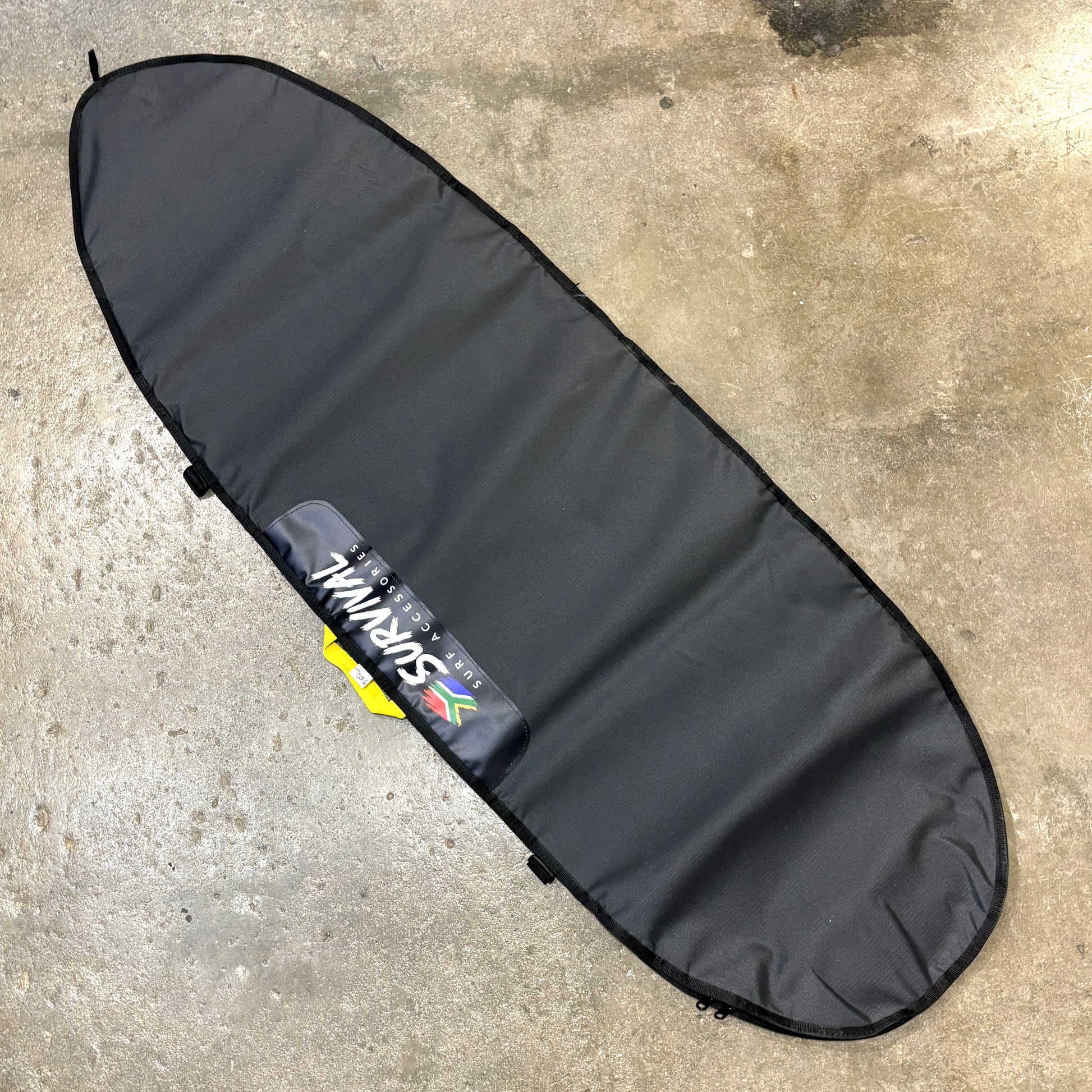 A black surfboard carrying bag laid diagonally on a concrete floor, featuring a visible Survival brand patch.
