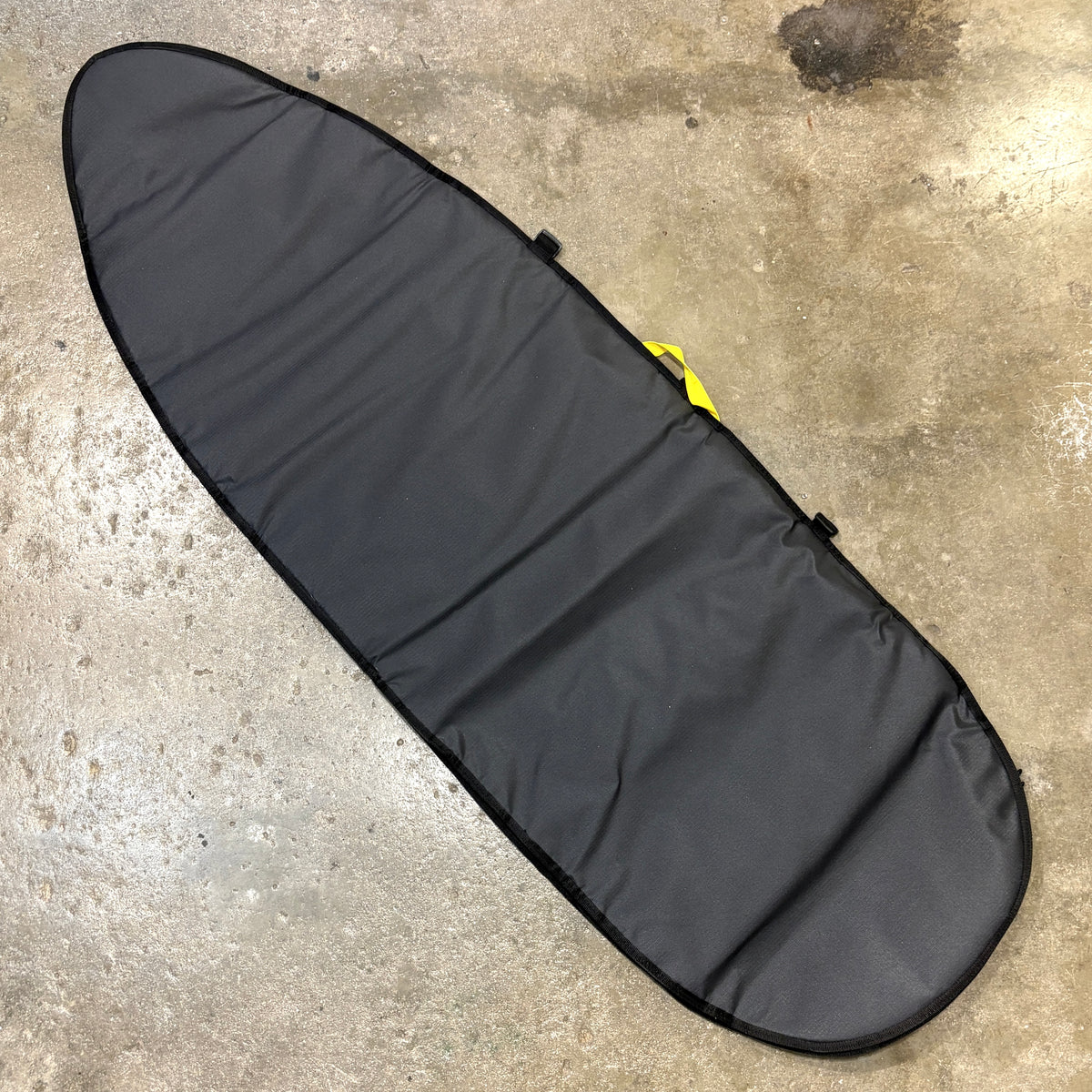A black surfboard carrying bag laid diagonally on a concrete floor, with a small yellow loop visible near the upper right side.