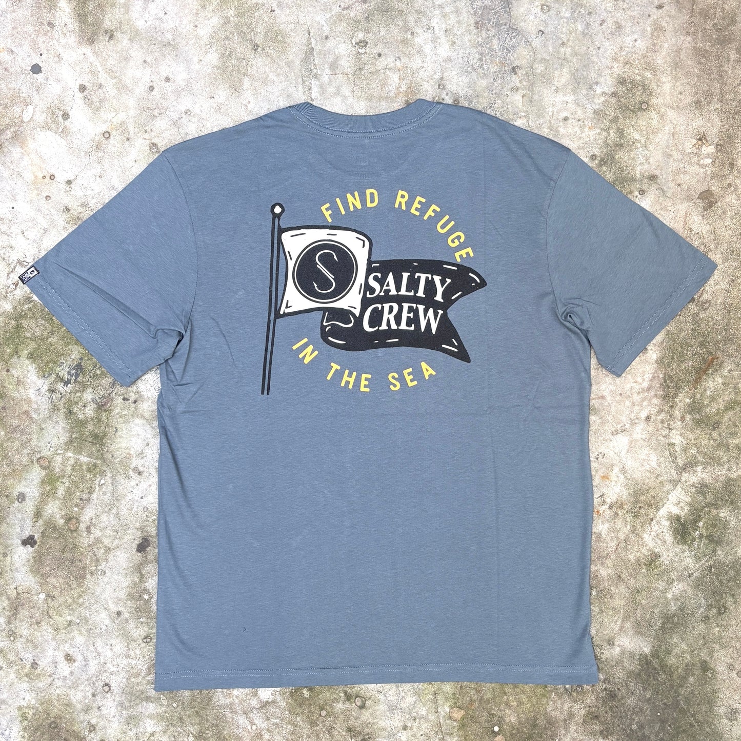 A heather slate blue t-shirt laid flat on a textured concrete surface, showing the back with a graphic print that reads 'FIND REFUGE IN THE SEA' and 'SALTY CREW' on a flag design.