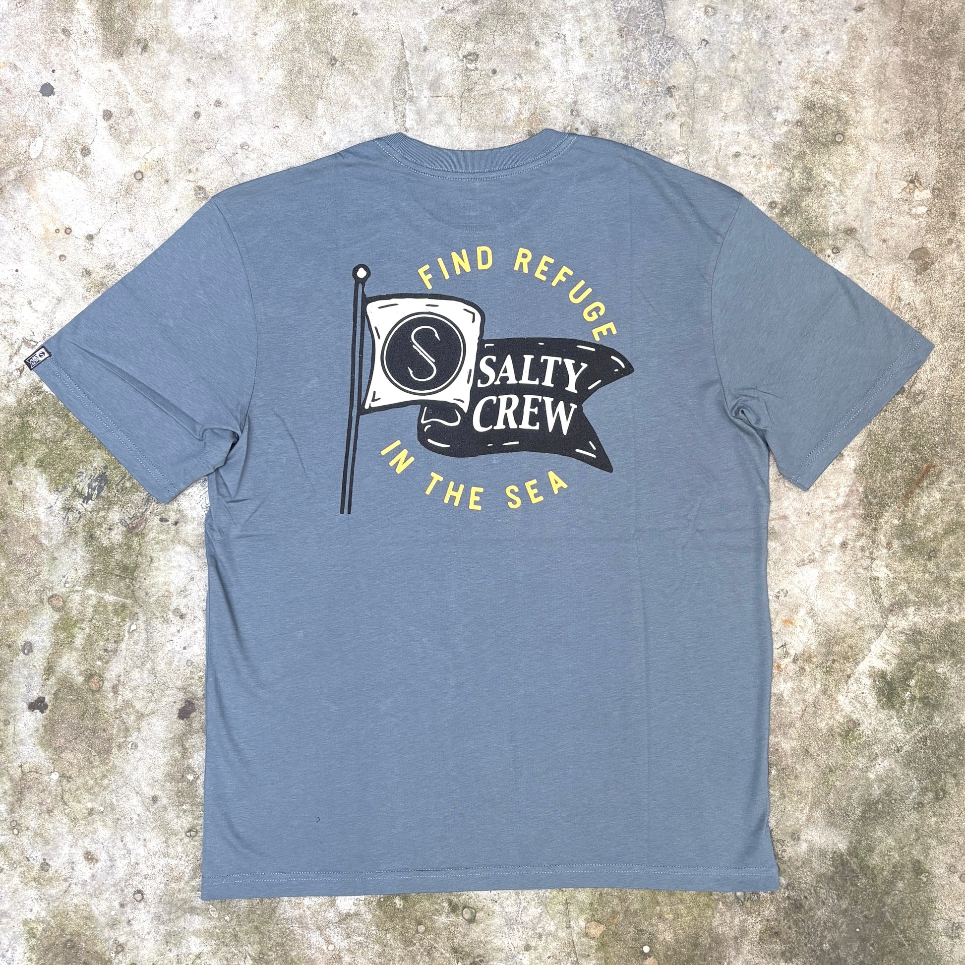 A heather slate blue t-shirt laid flat on a textured concrete surface, showing the back with a graphic print that reads 'FIND REFUGE IN THE SEA' and 'SALTY CREW' on a flag design.